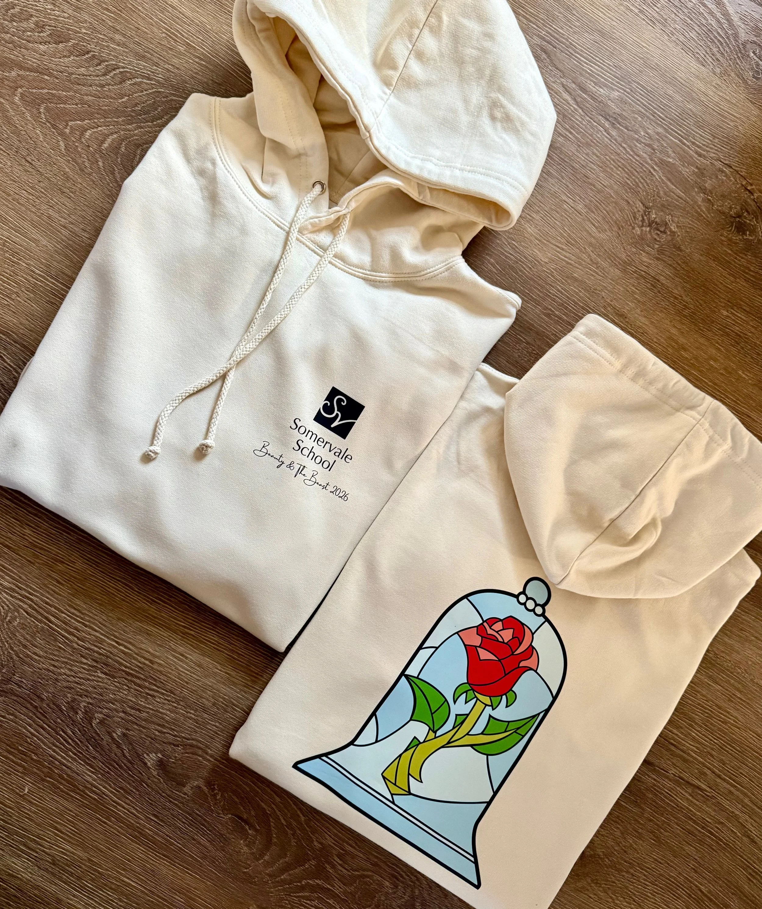 Two cream-colored hoodies with graphics and text, folded and laid on a wooden surface. One hoodie features a school logo and text; the other has a stained-glass style illustration of a red rose inside a blue-tinted bell shape.