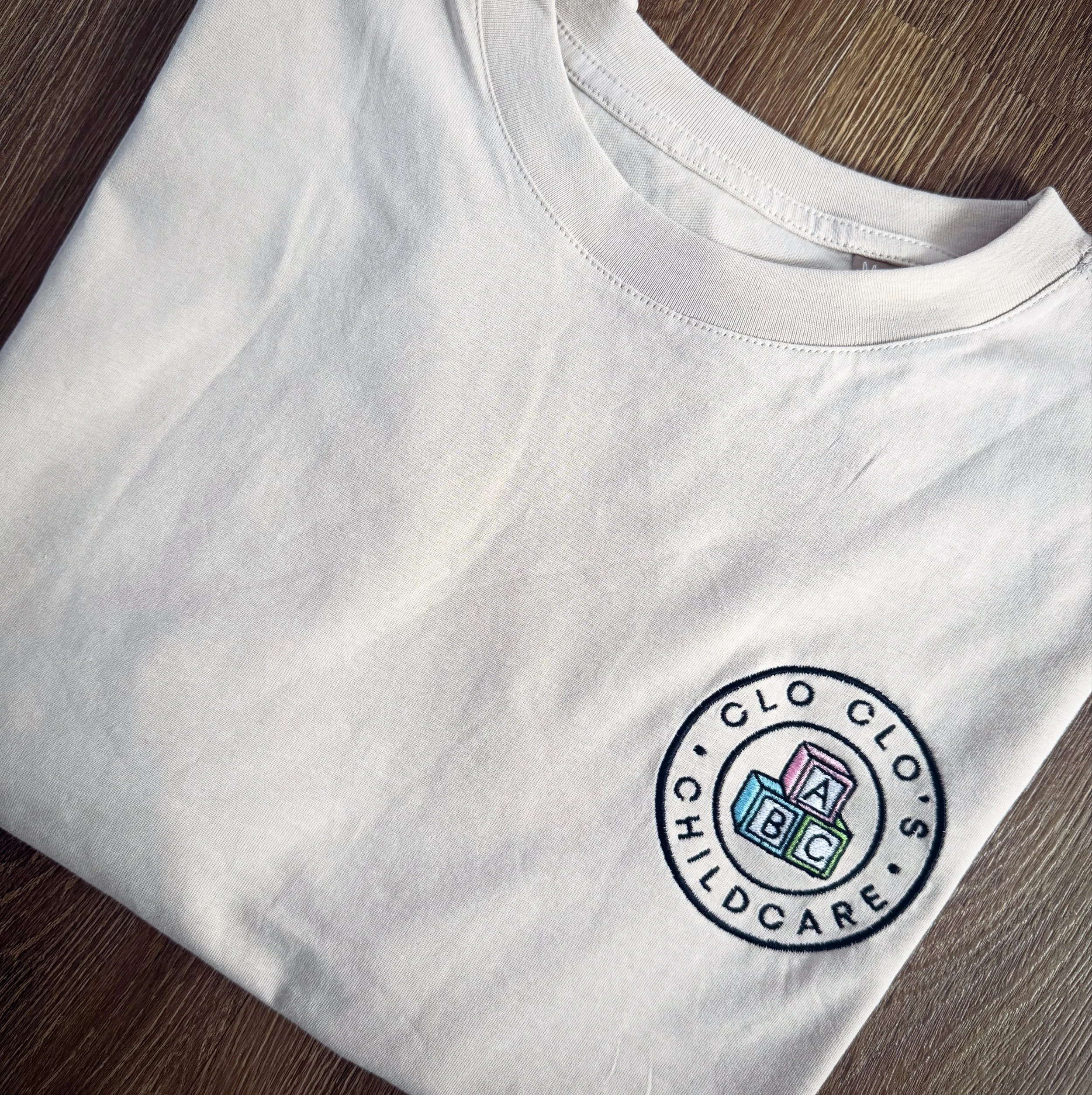 A white t-shirt with an embroidered logo of a childcare center reading 'CLO CLO CHILDCARE' with colorful blocks displaying 'A B C' on the chest.