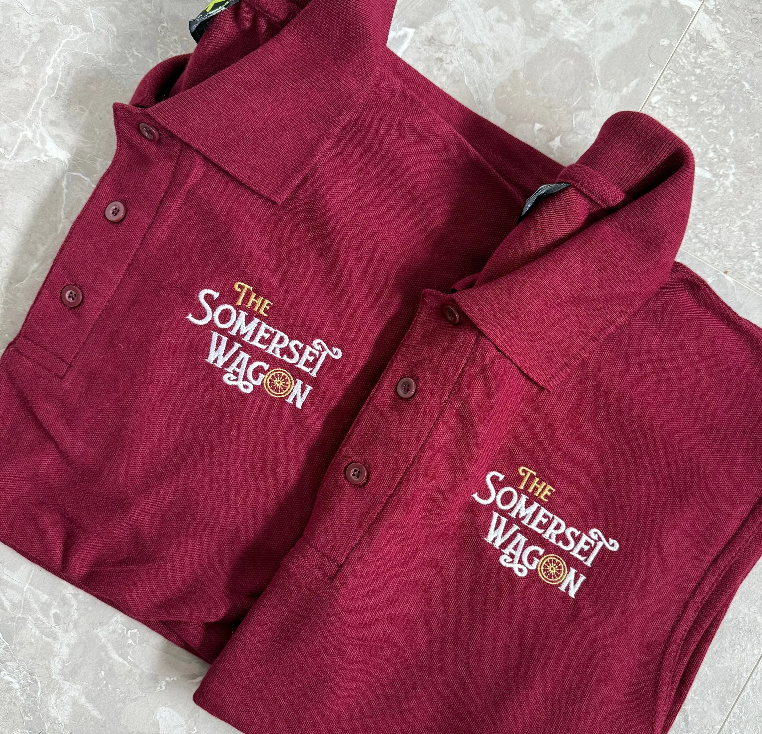 Two maroon polo shirts with embroidery reading "The Somerset Wagon" and a wheel emblem, folded and placed on a gray tile floor.