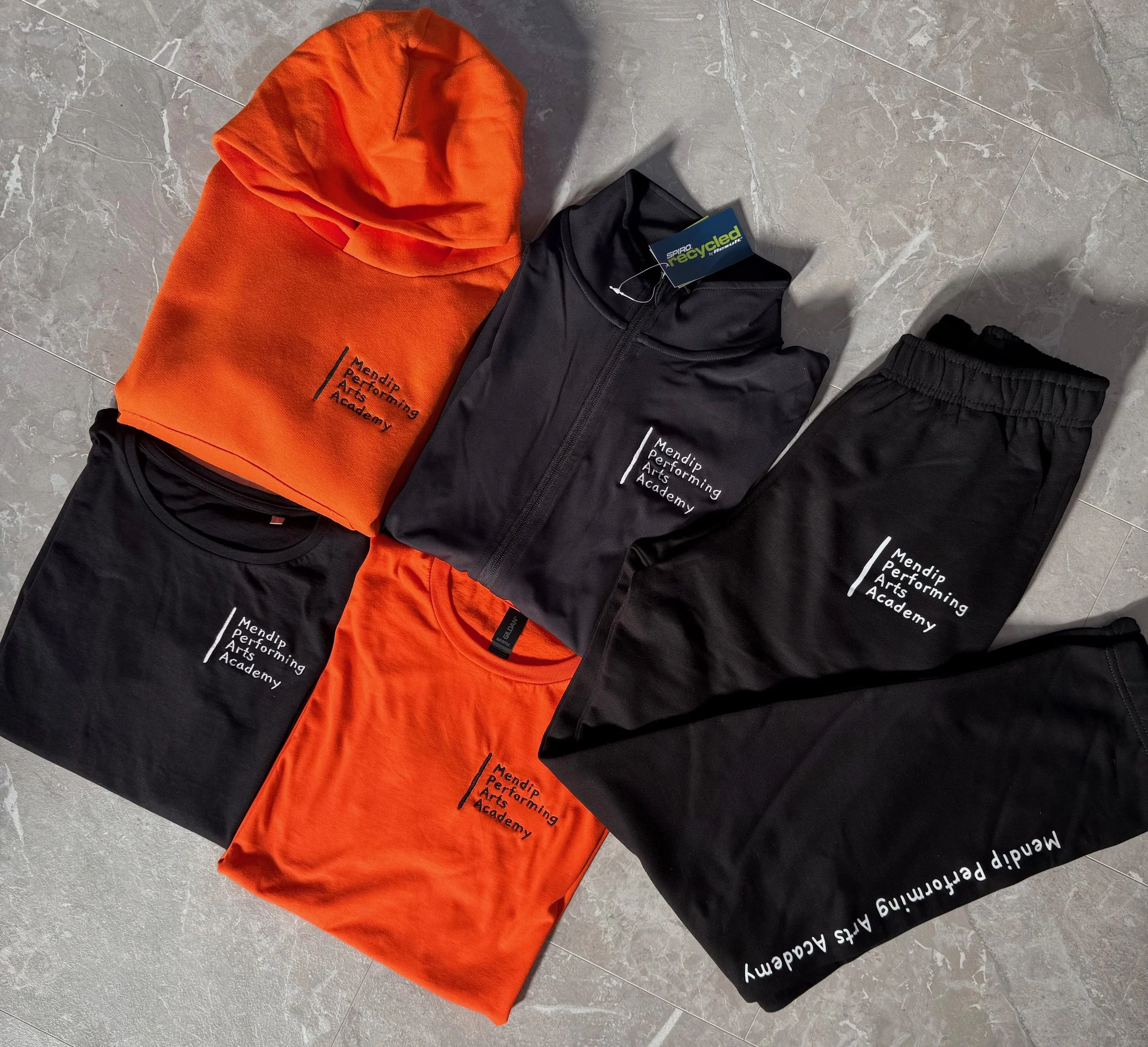 Arranged clothing items on a gray tile floor, including two orange hoodies, two black t-shirts, one black zip-up jacket, and black pants, all with 'Mendip Performing Arts Academy' embroidered on them.