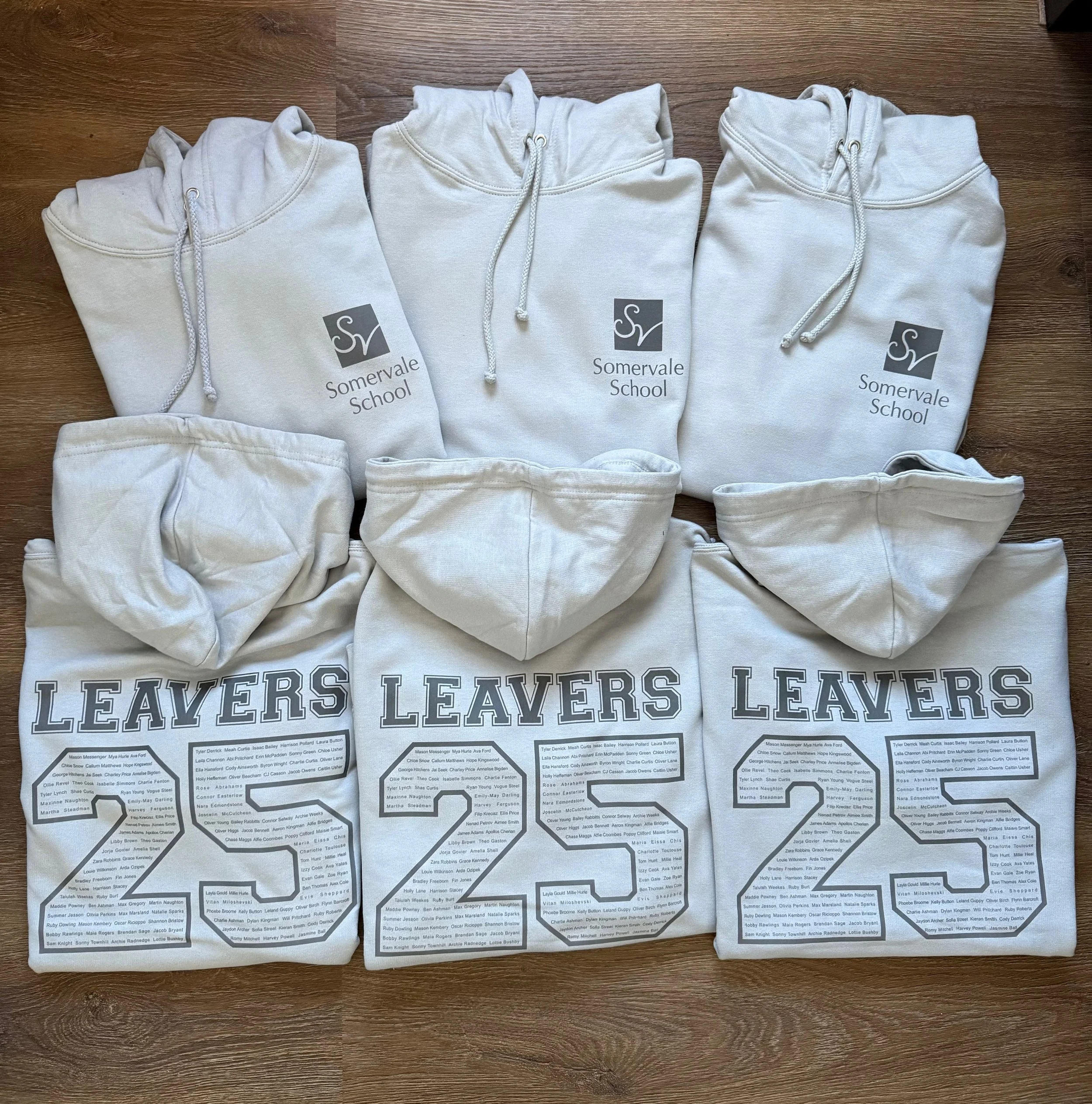 Six white hoodies with printed logos and text laid out on a wooden floor. The top row has three hoodies with 'Somervale School' logos, while the bottom row has three hoodies with 'Leavers 2023' text and large numbers, each with entered names inside.