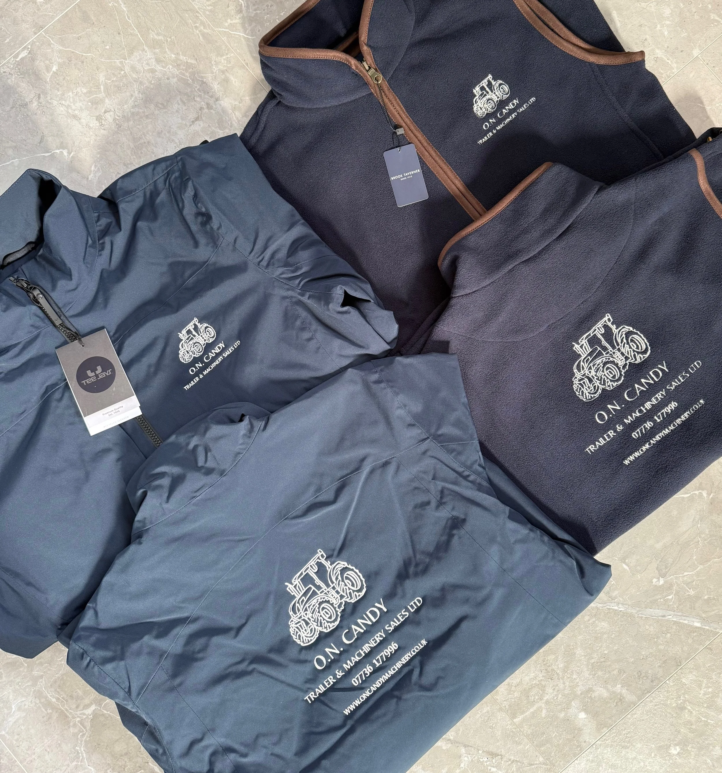 Three jackets with embroidered logos and text for O.N. CANDY trailer and machinery sales, displayed on a marble floor.