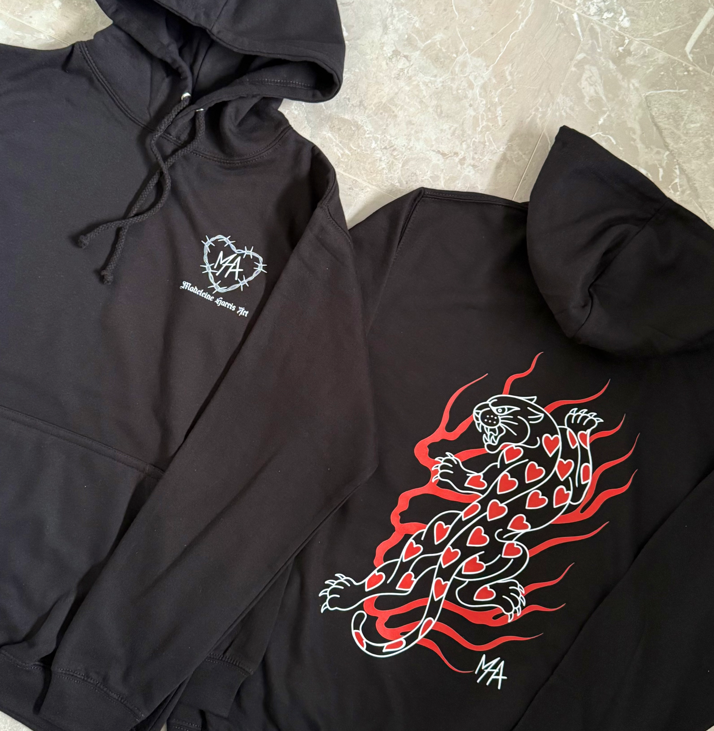 Two black hoodies, one with a white logo and text reading 'Madeleine Harris '19' and initials 'MA', the other featuring a colorful graphic of a roaring tiger with red hearts and flames, and initials 'MA'