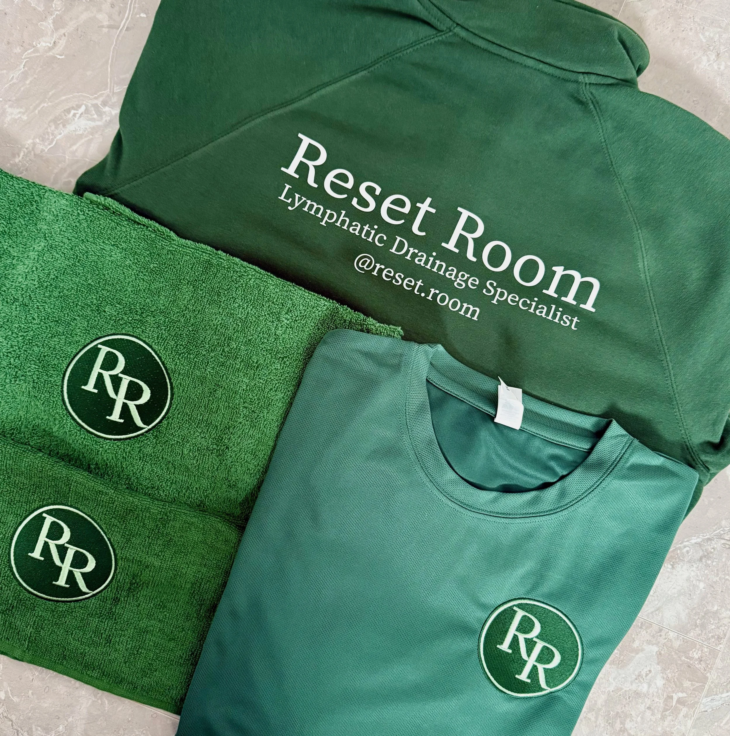 A green hoodie with white text that reads 'Reset Room Lymphatic Drainage Specialist @reset.room', a green towel with a circular logo featuring the initials 'RR' in white, and a green T-shirt with the same circular logo.