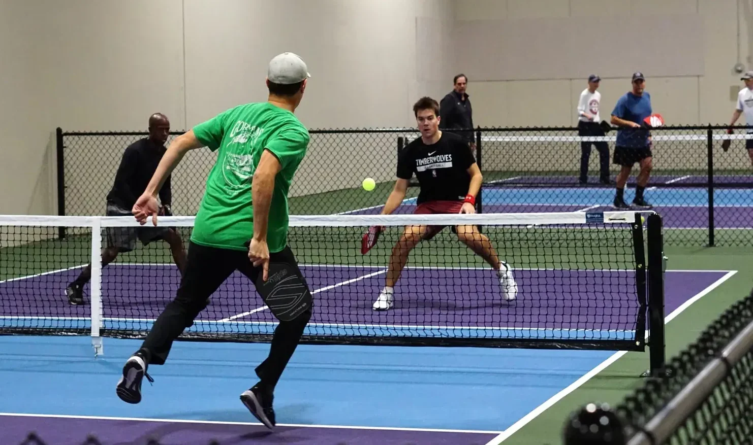 Two men playing pickleball indoors, with one about to hit the ball and the other ready to return, while three people watch in the background.
