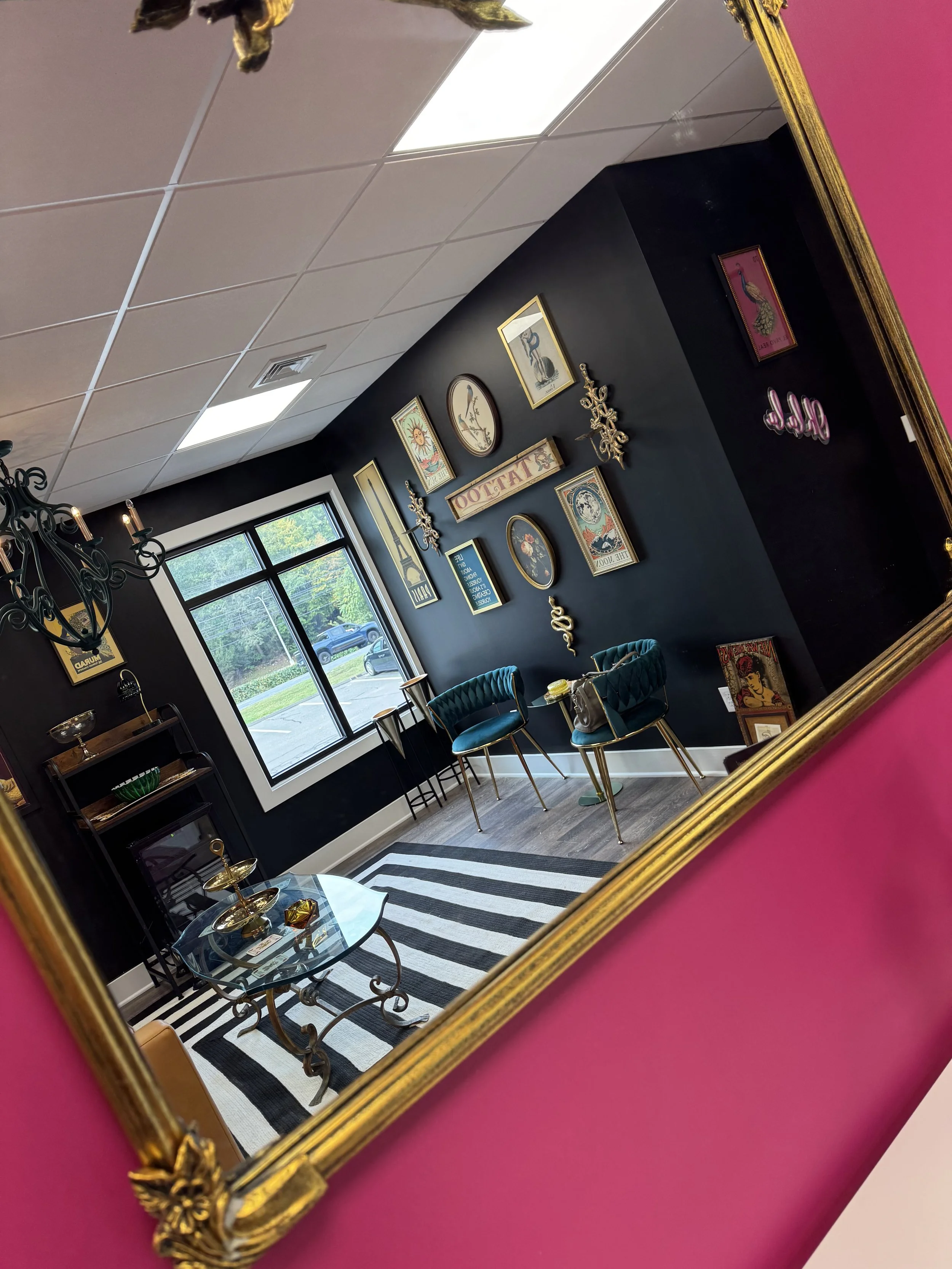 A decorated interior room with a black accent wall, framed artwork, teal velvet chairs, a glass coffee table with golden decorative items, a striped black and white rug, and a window showing greenery outside, all reflected in a gold-framed mirror on a pink wall.