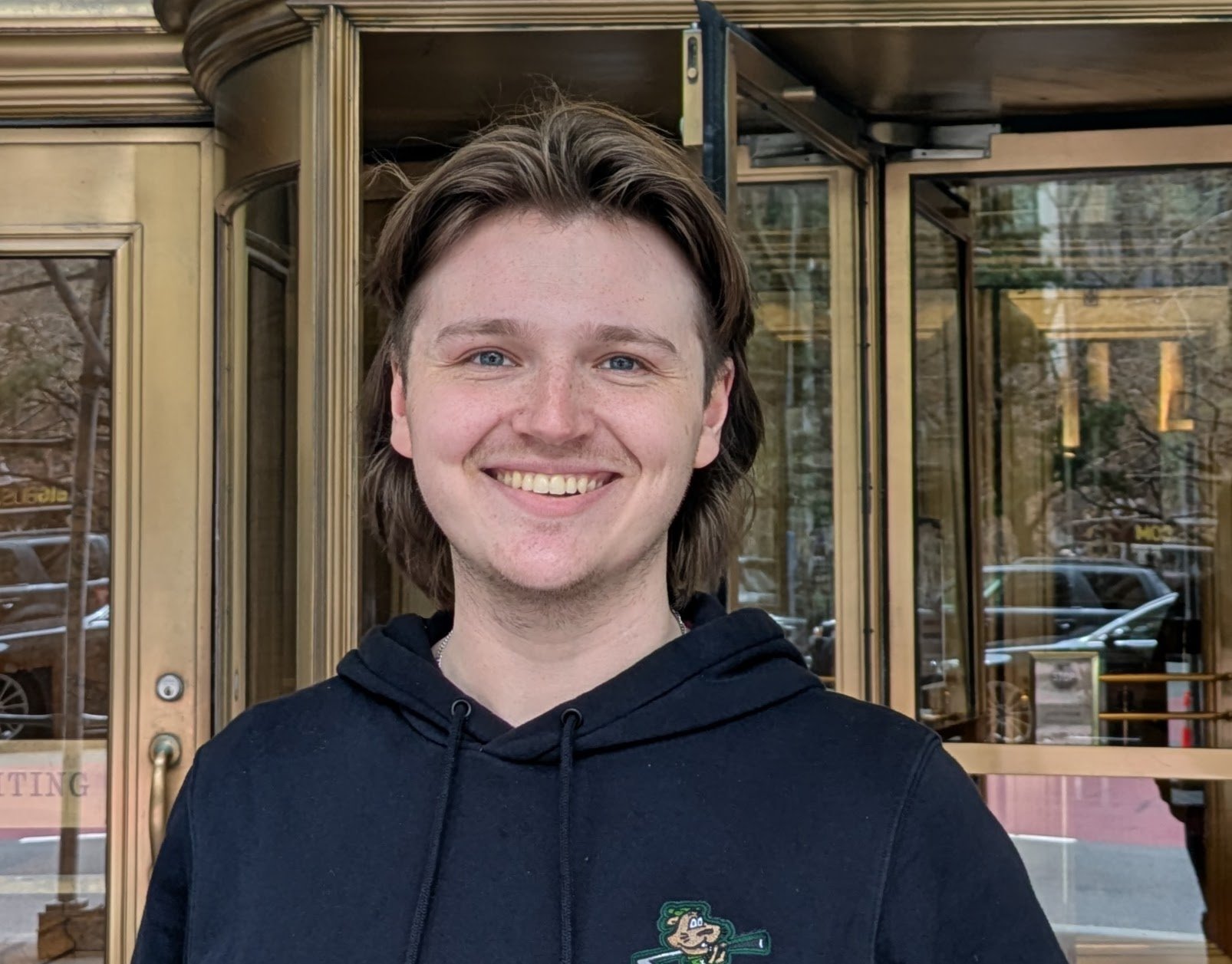 A young man with medium-length brown hair and blue eyes smiling in front of a glass door with a brass frame, reflecting trees and parked cars outside.