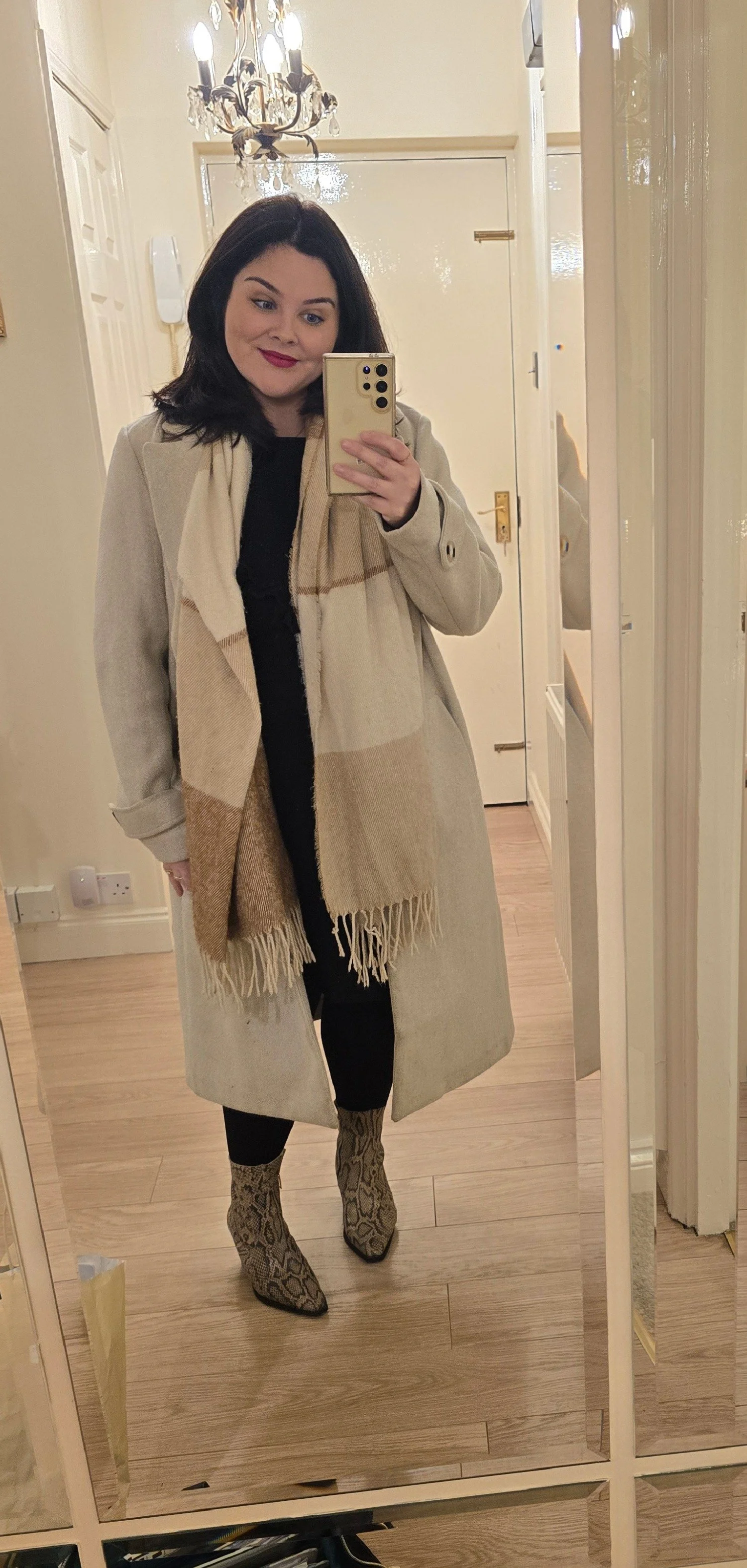 Woman with black hair taking a mirror selfie in a hallway, wearing a beige coat, matching scarf, black leggings, and snake print boots.