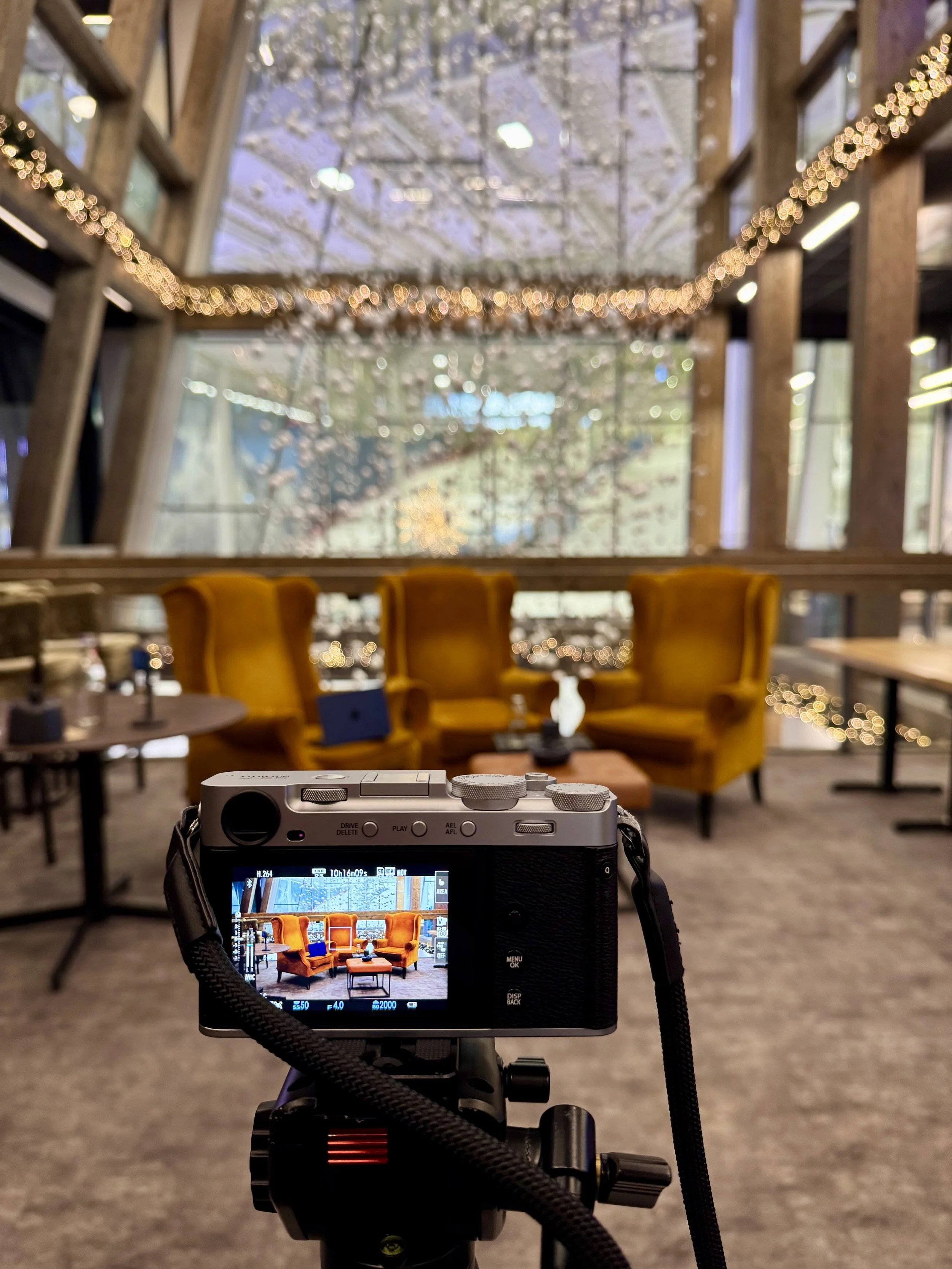 Et kamera på stativ som filmer en innendørsscene med sennepsgule lenestoler plassert foran et stort vindu dekorert med lysslynger og vinterdekorasjoner.