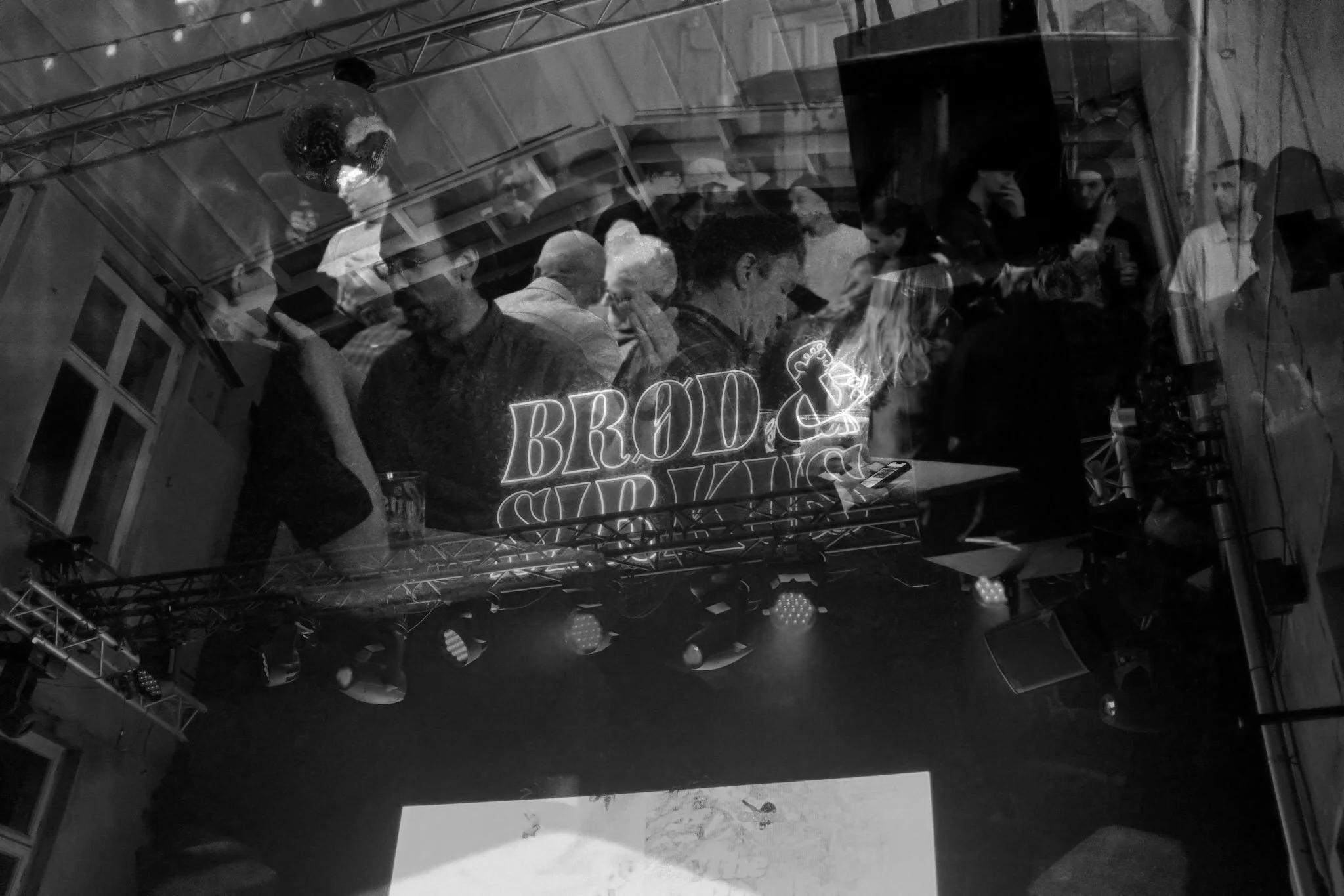A reflection of a crowded indoor event with people mingling, some looking at their phones, and a neon sign that reads 'BROD & SURF.' There are stage lights and equipment visible.