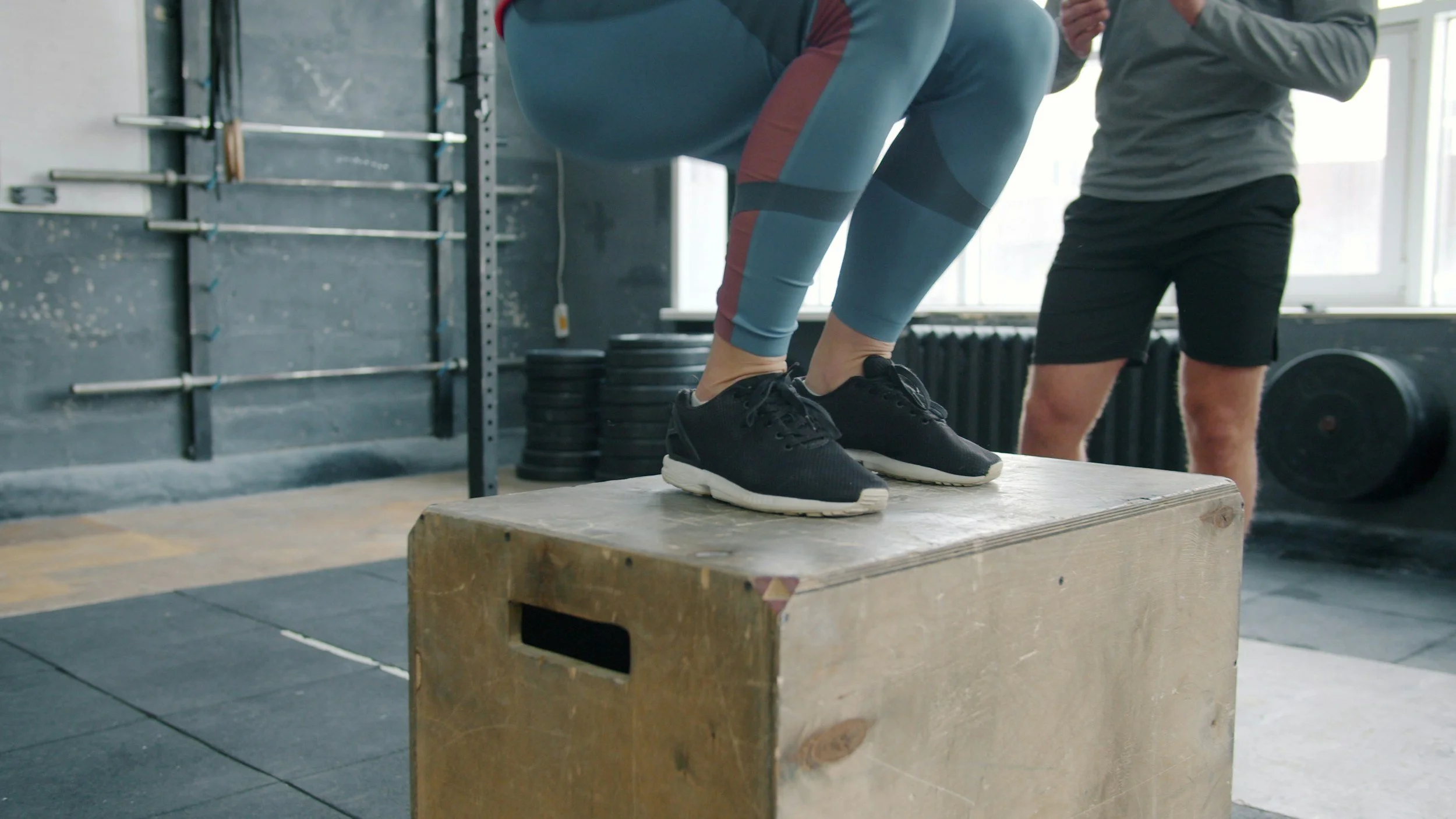Person performing box jump exercise on a wooden plyometric box at a gym, with a trainer standing nearby.