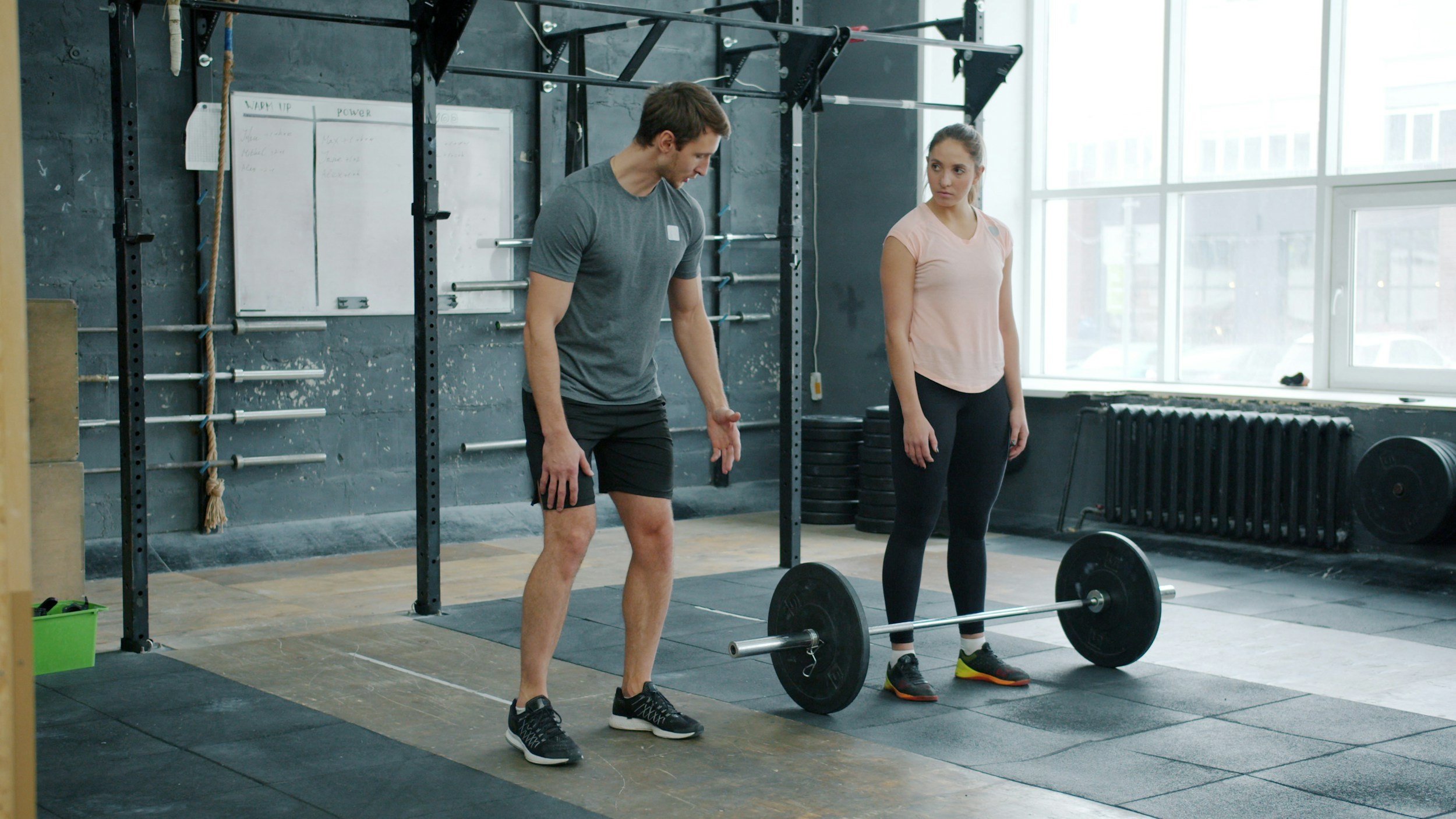 A man and a woman in a gym, preparing for a deadlift exercise, with the man demonstrating proper form and the woman watching.