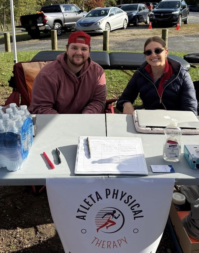 A man and a woman sit at a table outdoors with a banner that reads "Atleta Physical Therapy." The man is wearing a red cap backward and a maroon hoodie, and the woman is wearing sunglasses and a navy jacket. There are water bottles, hand sanitizer, a clipboard, and a laptop on the table.
