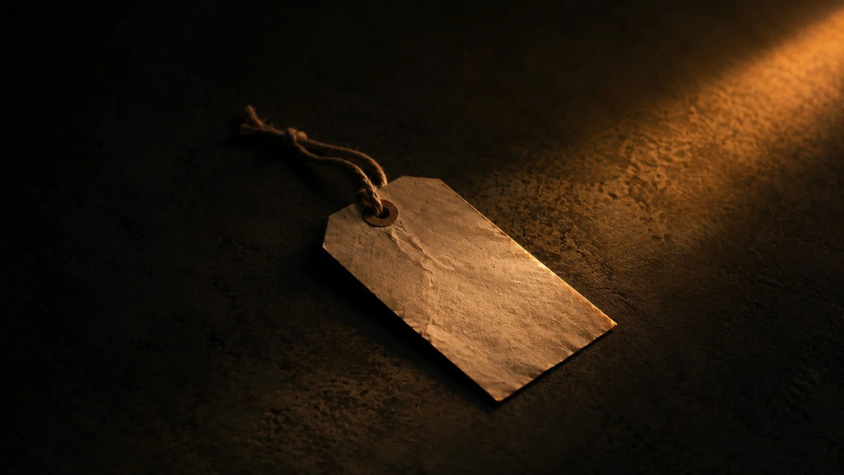 A blank, aged paper tag with a hole at the top, attached to a frayed string, lies on a dark surface illuminated by warm, low lighting.