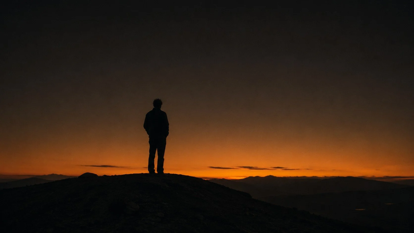 Silhouette of a person standing on a hilltop at sunset, with a colorful sky and distant mountains in the background.