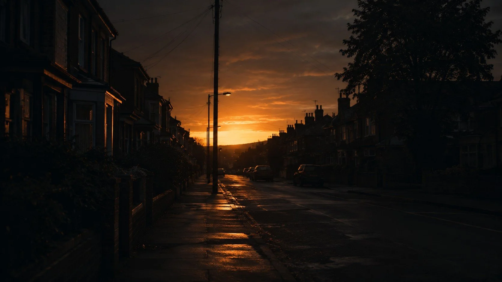 Sunset over a quiet residential street with wet pavement reflecting the golden sky, lined with dark houses and parked cars.