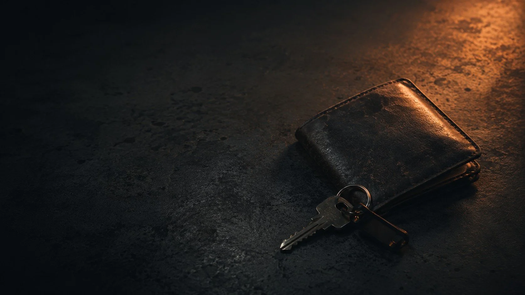 A black leather wallet with a set of keys attached, lying on a dark textured surface illuminated by warm lighting.  TASR CONSULTING HERO TAKE ACTION SEE RESULTS MEN SELF HELP.