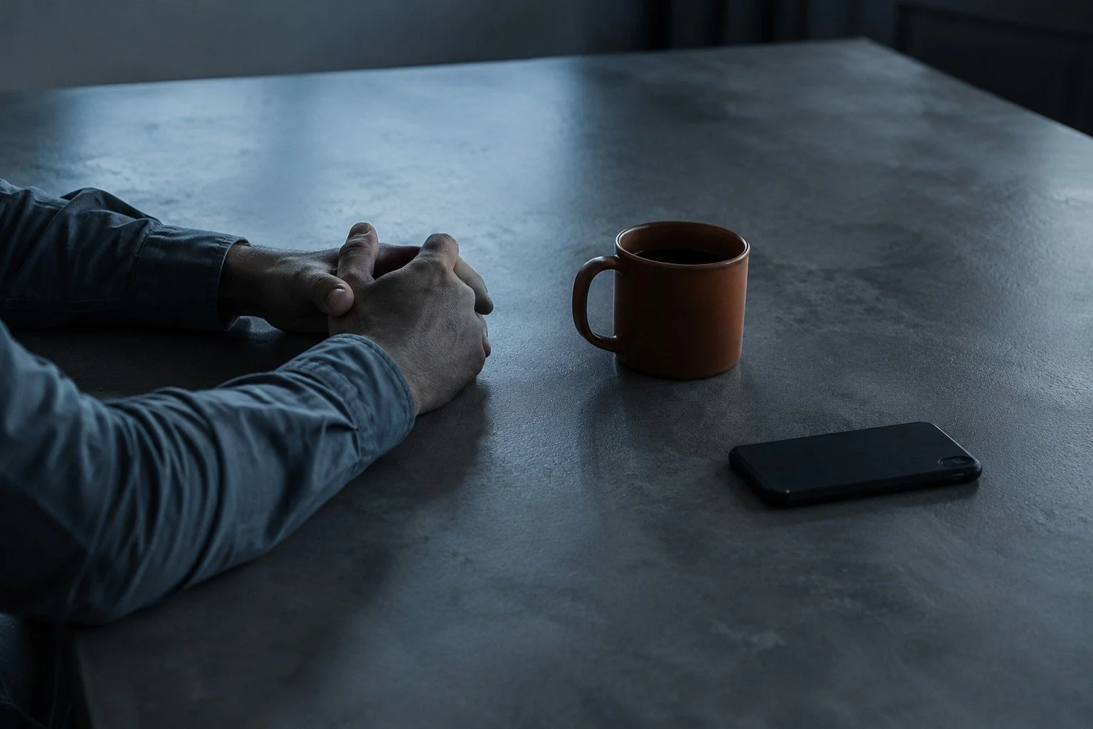 Person with hands clasped resting on a table next to a brown mug and a black smartphone.  TASR CONSULTING HERO TAKE ACTION SEE RESULTS MEN SELF HELP.