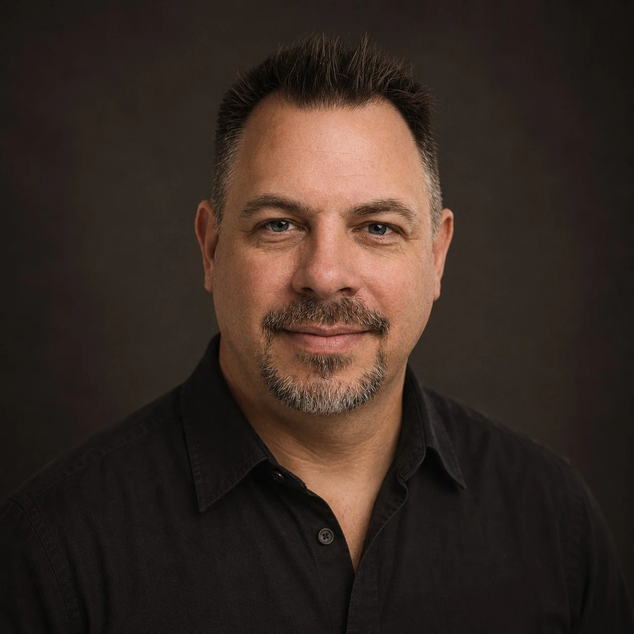 Headshot of a middle-aged man with short, dark, spiked hair, blue eyes, and a goatee, wearing a black button-up shirt against a dark background.