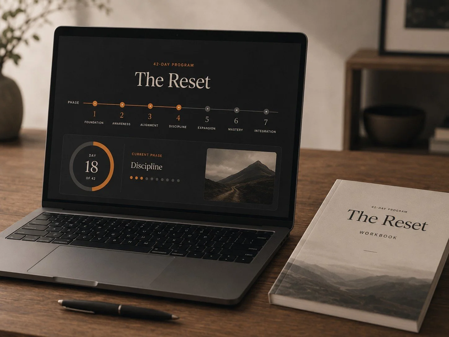 Laptop and workbook on a wooden desk showing a 42-day program called 'The Reset' with a focus on 'Discipline' on day 18. The program has seven phases from foundation to integration. There is a pen in front of the laptop and a mountain landscape picture on the screen.