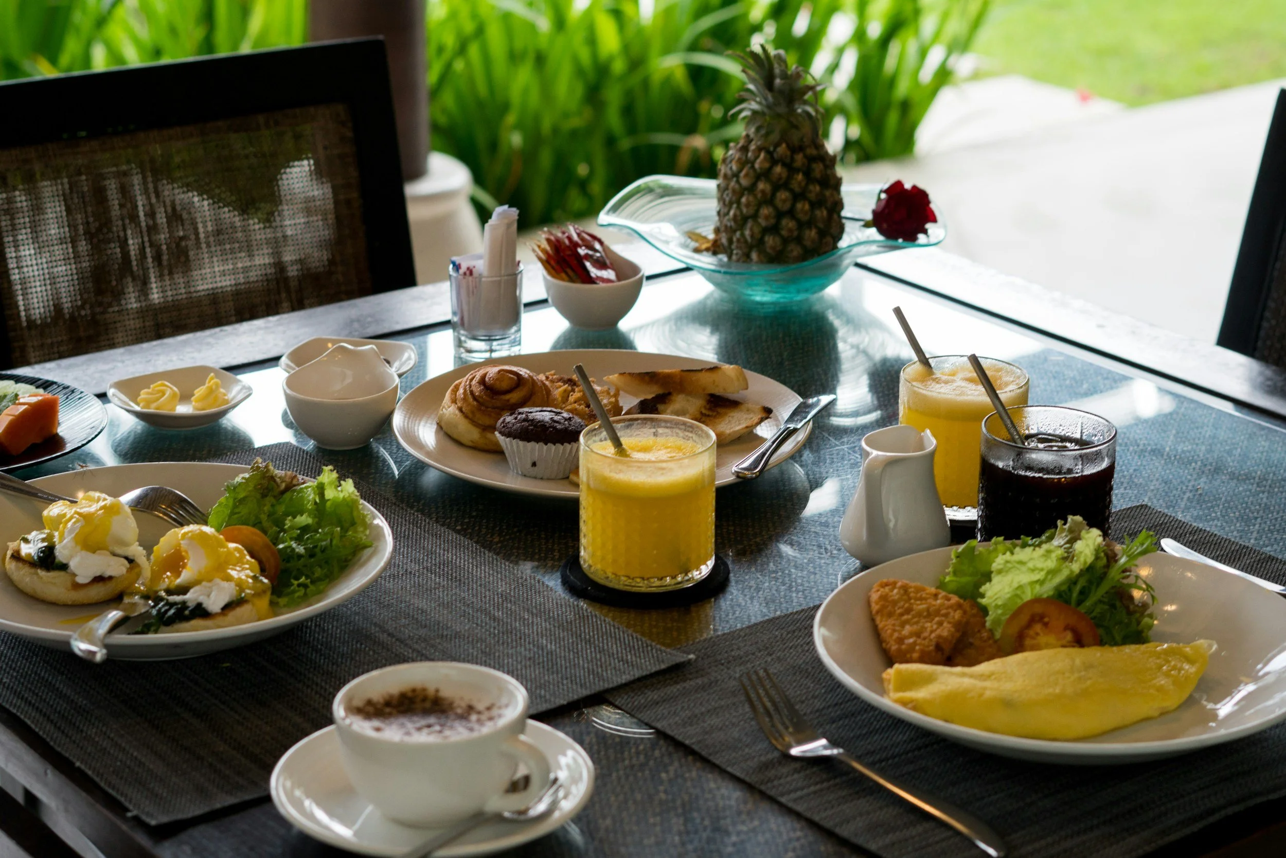 Breakfast table with assorted dishes including eggs benedict, salad, pastries, fruit, orange juice, and coffee, set near a window with greenery outside.