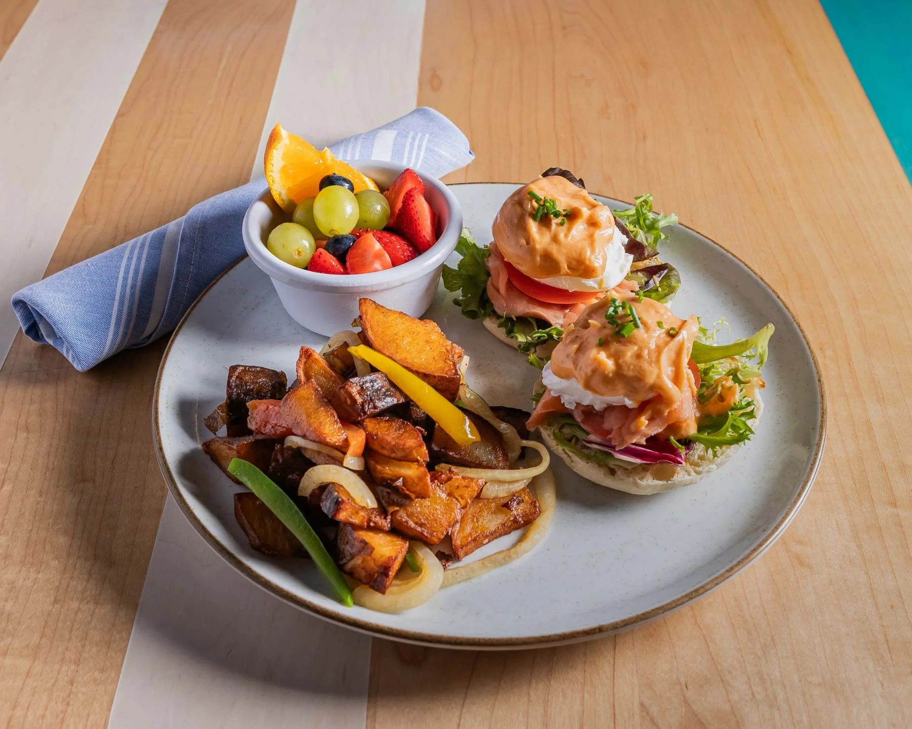 Plate with two open-faced sandwiches topped with smoked salmon, poached eggs, and hollandaise sauce, a side of roasted vegetables, and a small bowl of mixed fruit.