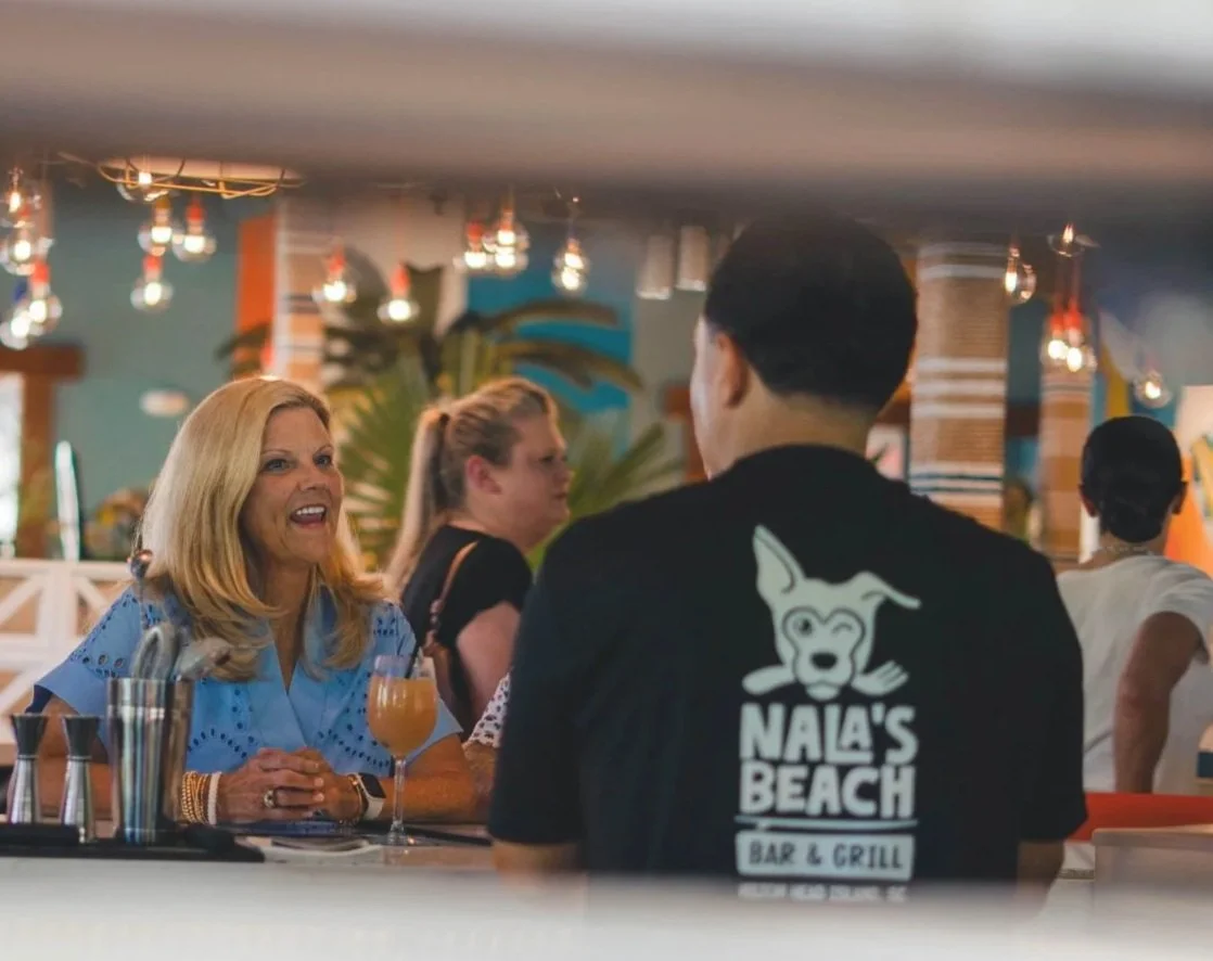 A woman with blonde hair smiling and talking at a bar in a restaurant or bar setting. She has a drink in front of her. The background shows other patrons and tropical decor.