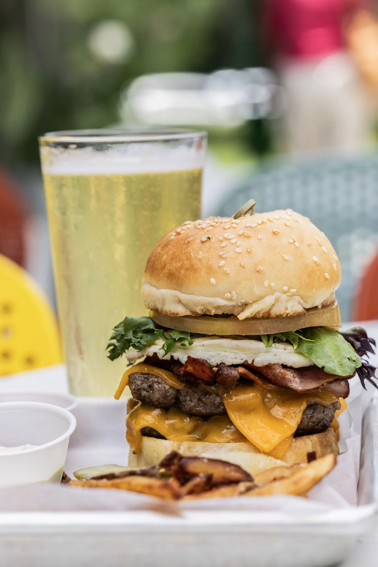 A tall glass of beer behind a large cheeseburger with lettuce, tomato, cheese, bacon, and a sesame seed bun, served with fries and a small dipping cup of sauce.