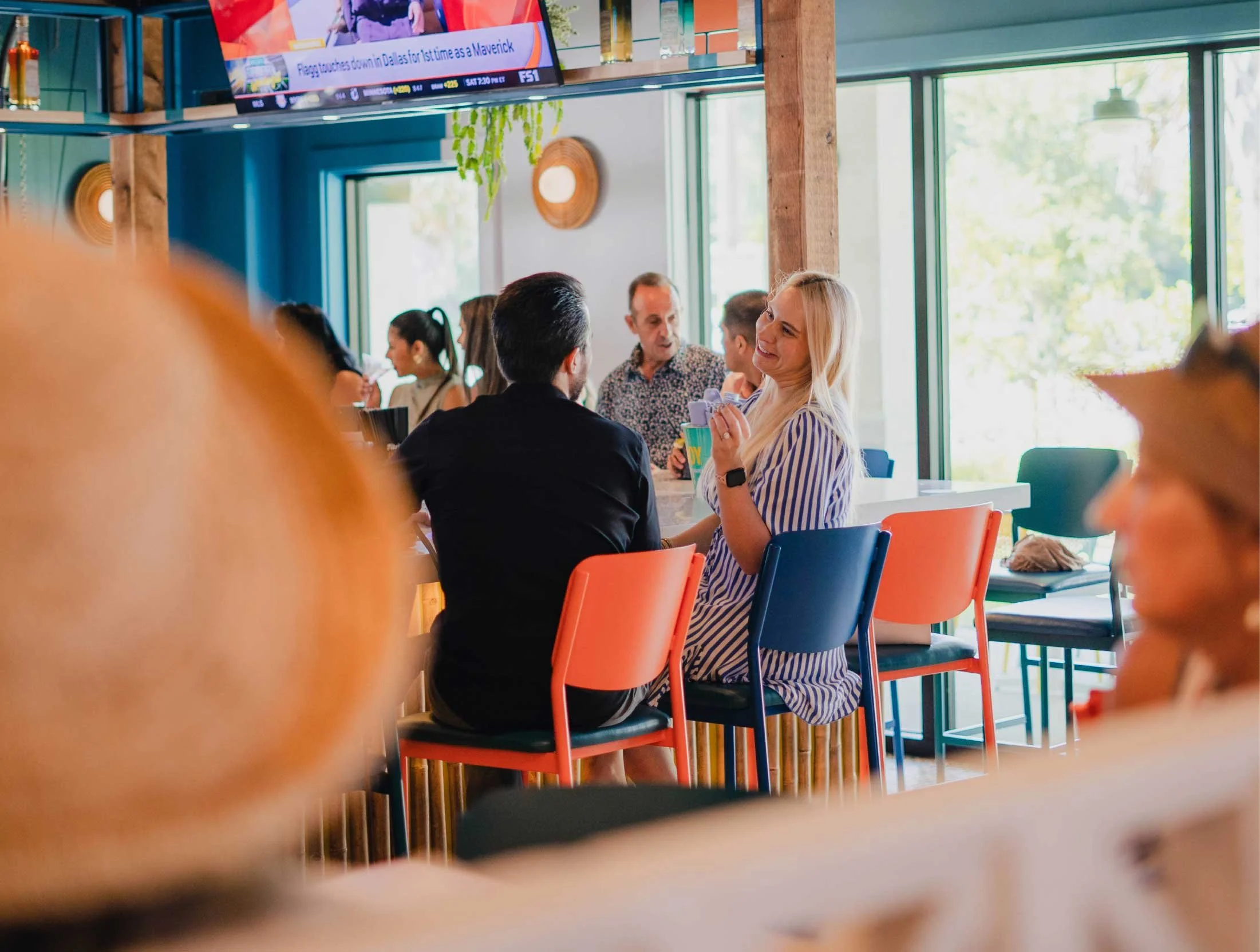 People sitting at a colorful cafe, engaging in conversations, with large windows and a television screen visible in the background.