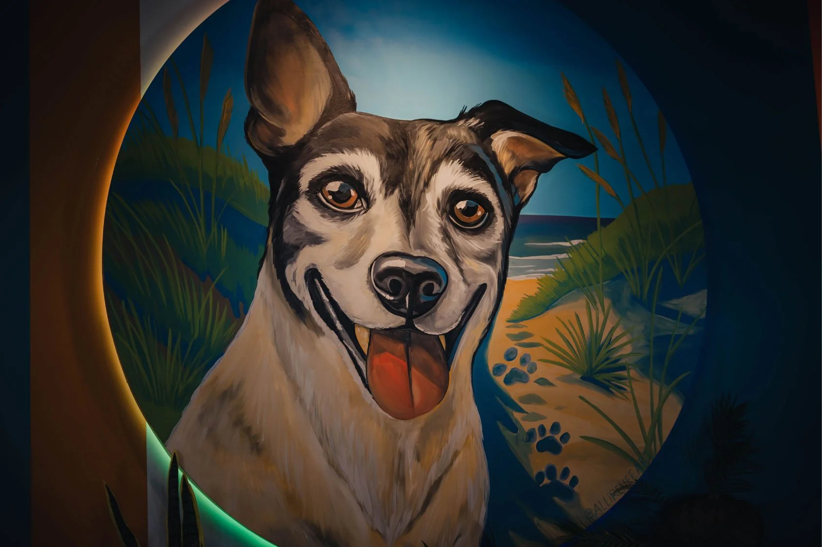 Colorful mural of an Australian Shepherd dog with a happy expression, set against a beach scene with sand, plants, and ocean in the background.