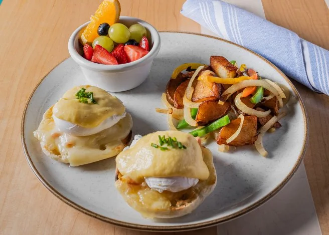 Plate with eggs Benedict, a side of breakfast potatoes topped with onions and bell peppers, and a small bowl of mixed fruit.