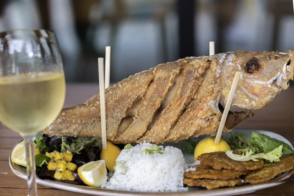A plate with a fried fish, rice, salad, lemon wedges, and plantains, with a glass of white wine in the foreground.