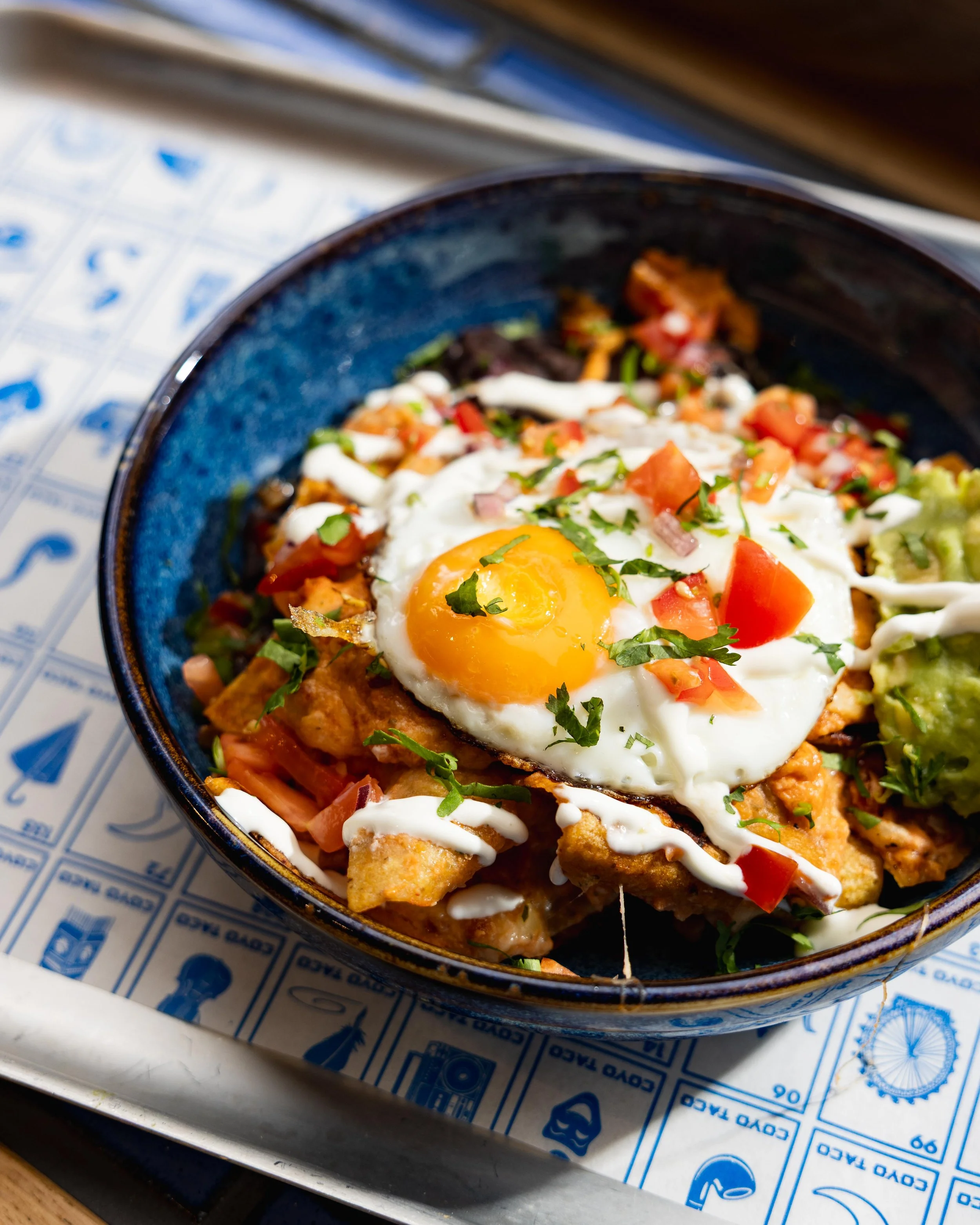 Close-up of a bowl of Mexican chilaquiles topped with a fried egg, chopped tomatoes, green onions, cilantro, sour cream, and sliced avocado on the side, placed on a tray with blue and white printed paper.
