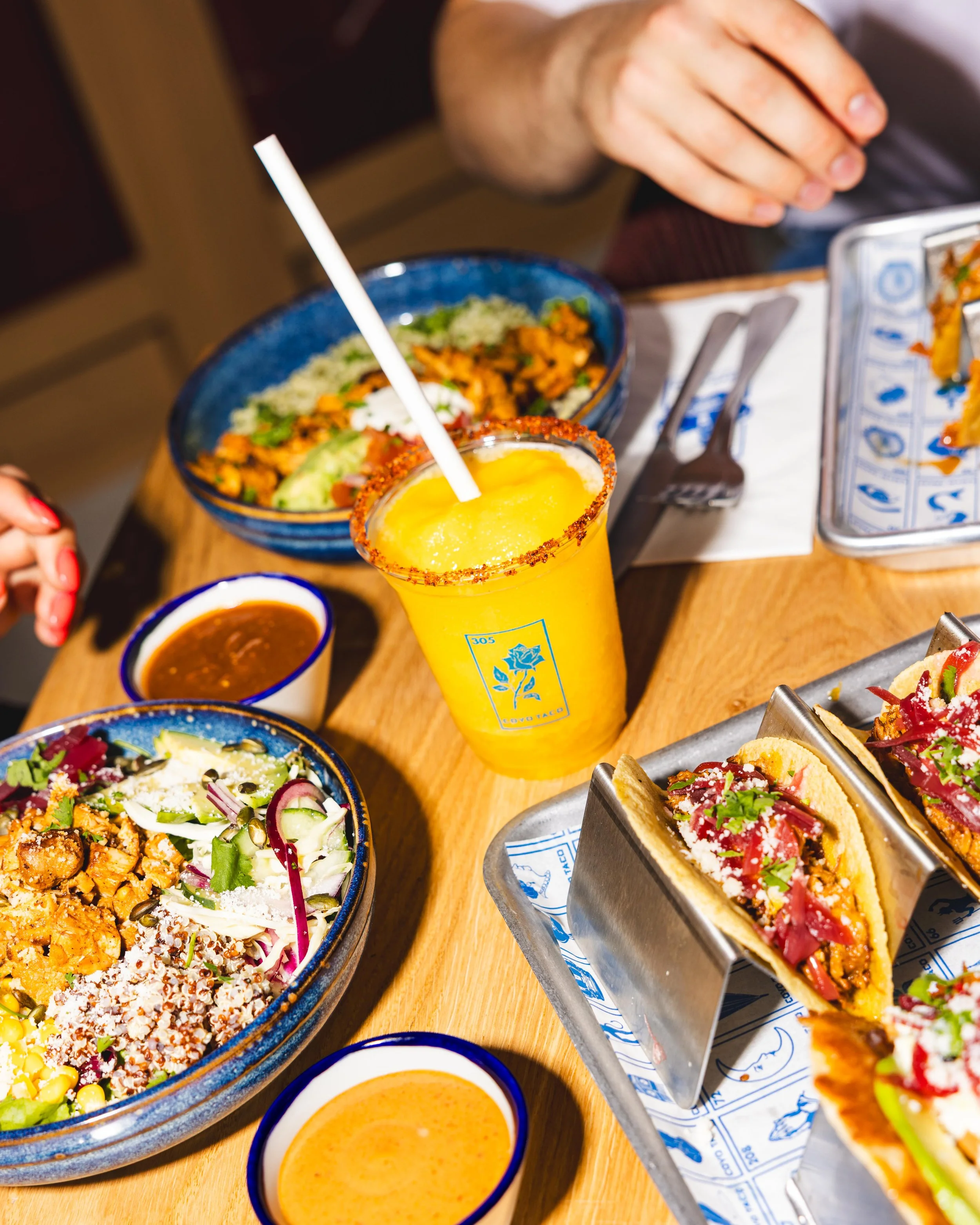 Mexican food and drinks on a wooden table, including tacos, salads, salsa, and a yellow beverage with a straw and chili powder rim.
