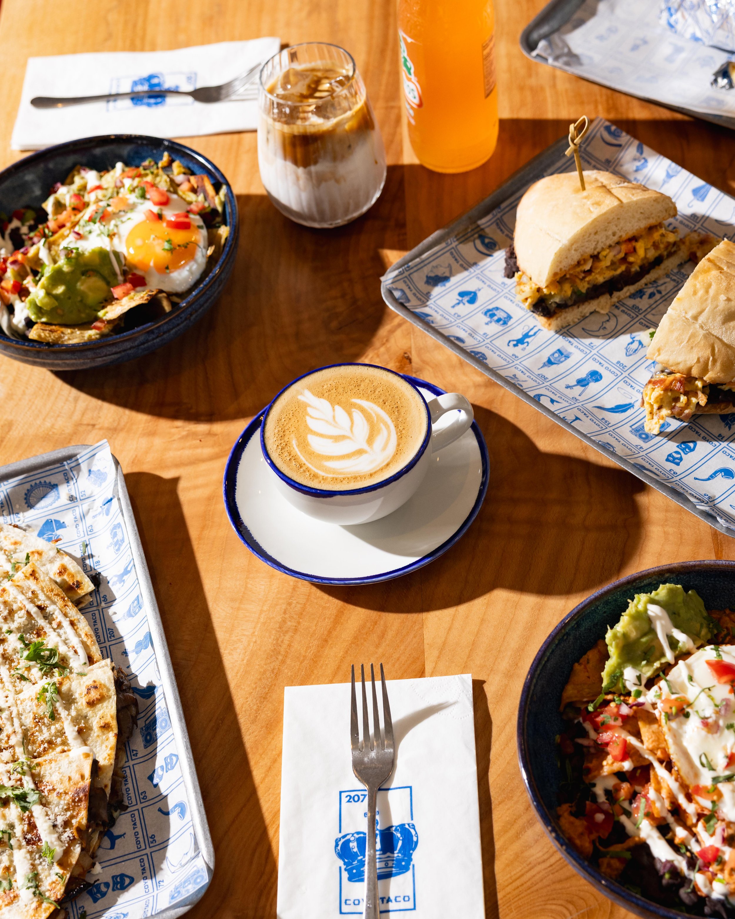 Table with various Mexican dishes, including a taco with meat and vegetables, a bowl of Mexican rice, a drink with ice and cream, a bottle of orange soda, and a latte with latte art, with utensils and napkins on a wooden table.