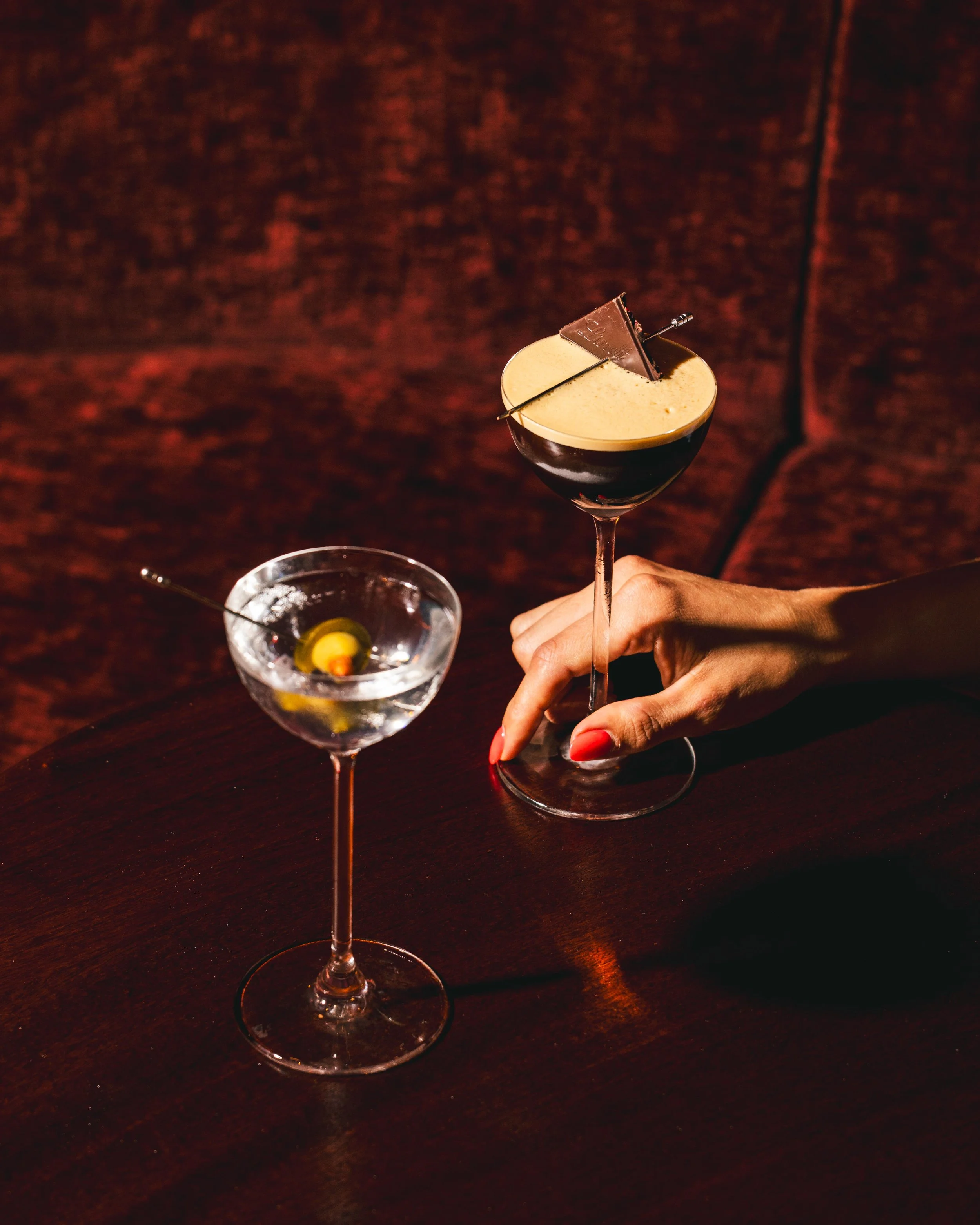 A hand holding a martini glass with a creamy cocktail garnished with a piece of chocolate and a small metallic decorative pick, with a separate glass containing ice and a lemon twist on a dark wooden table.