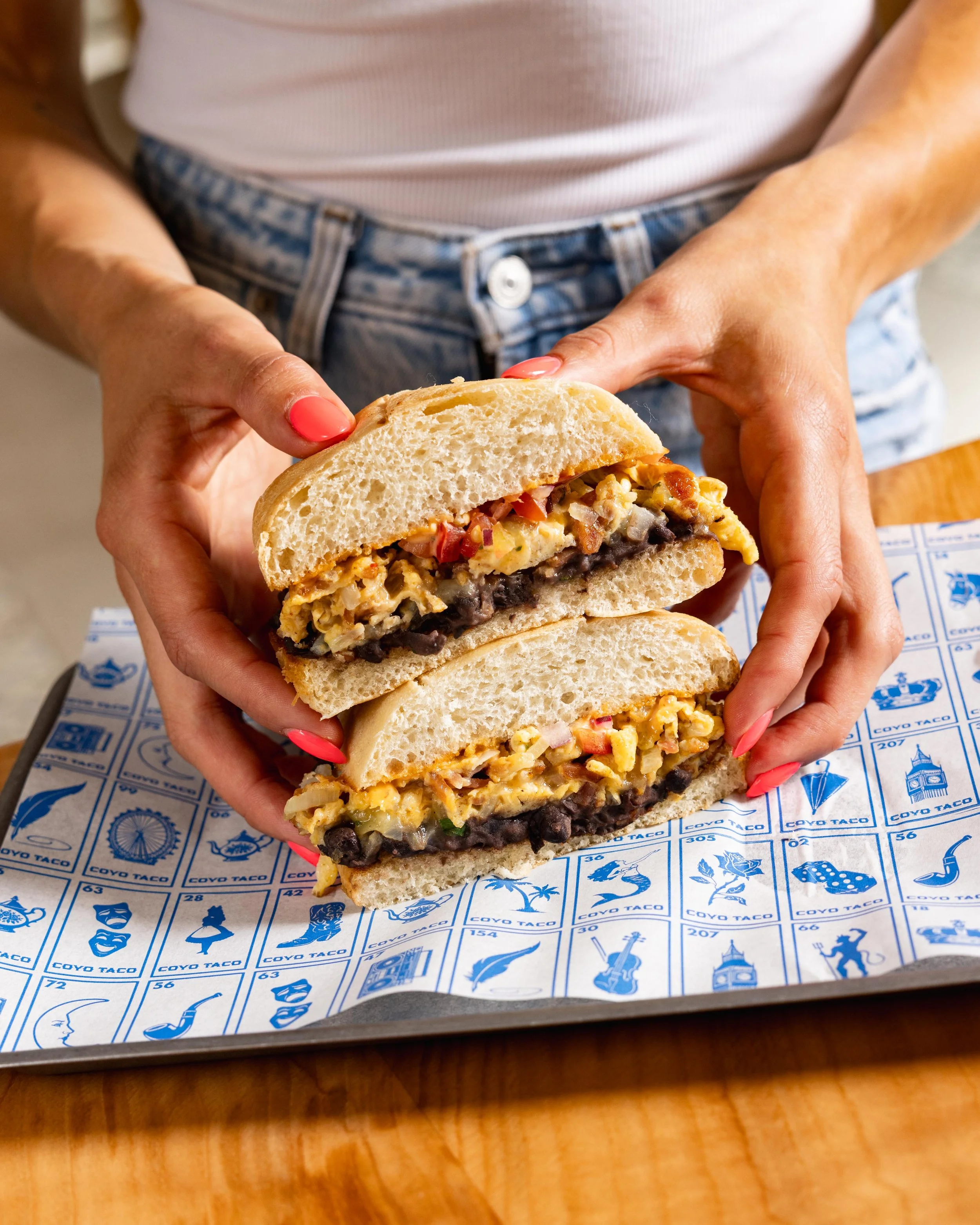 Close-up of a sandwich being held by a person with red nail polish. The sandwich has a filling of eggs, tomatoes, black beans, and other ingredients inside a bread roll. The person is wearing a white shirt and denim shorts. The sandwich is on blue and white paper with various illustrations and text from Coyo Taco, placed on a wooden table.