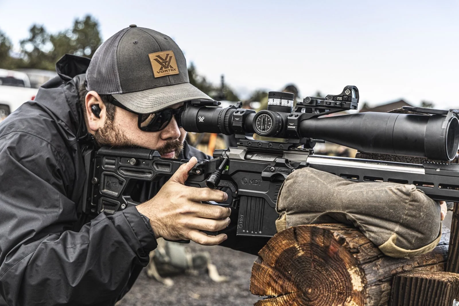 Man aiming a rifle with a scope, wearing sunglasses, a gray baseball cap, and a black jacket, leaning on a wooden log.