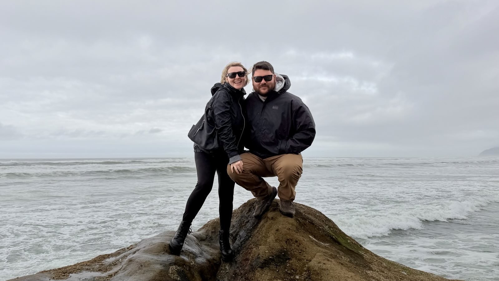A woman and a man posing on a large rock at the beach, wearing black jackets and sunglasses, with the ocean and cloudy sky in the background.