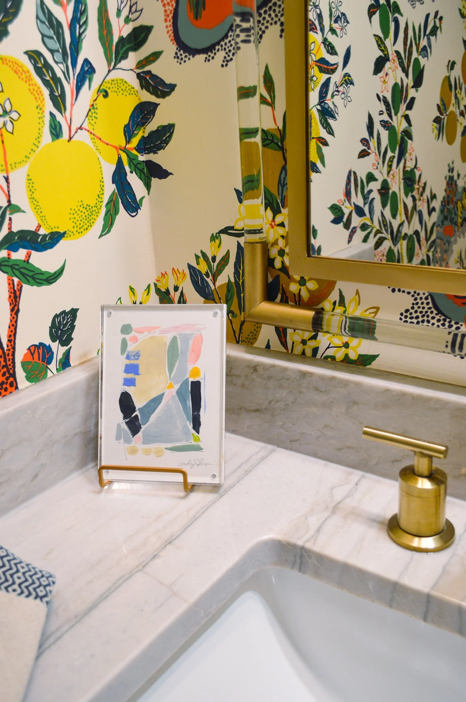 A bathroom countertop with a colorful abstract art piece on a stand, a gold faucet, and vibrant, botanical-patterned wallpaper and mirror reflecting the wallpaper.