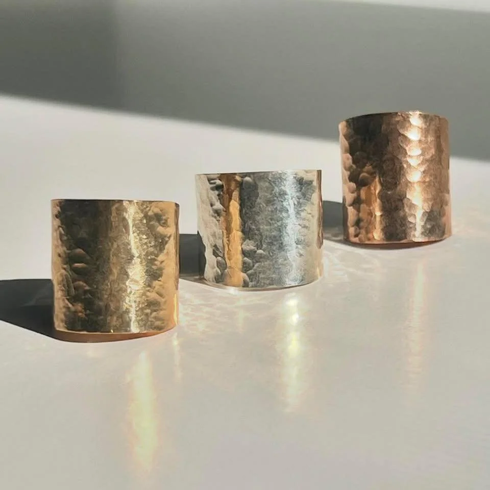 Three hammered metal rings, two gold-colored and one silver-colored, arranged in a row on a white surface with shadows and light reflections.