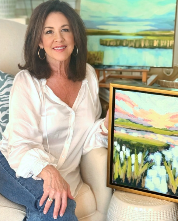 A woman with shoulder-length brown hair, wearing a white blouse, sitting on a beige couch next to a framed landscape painting of a marshland with water and pink clouds.
