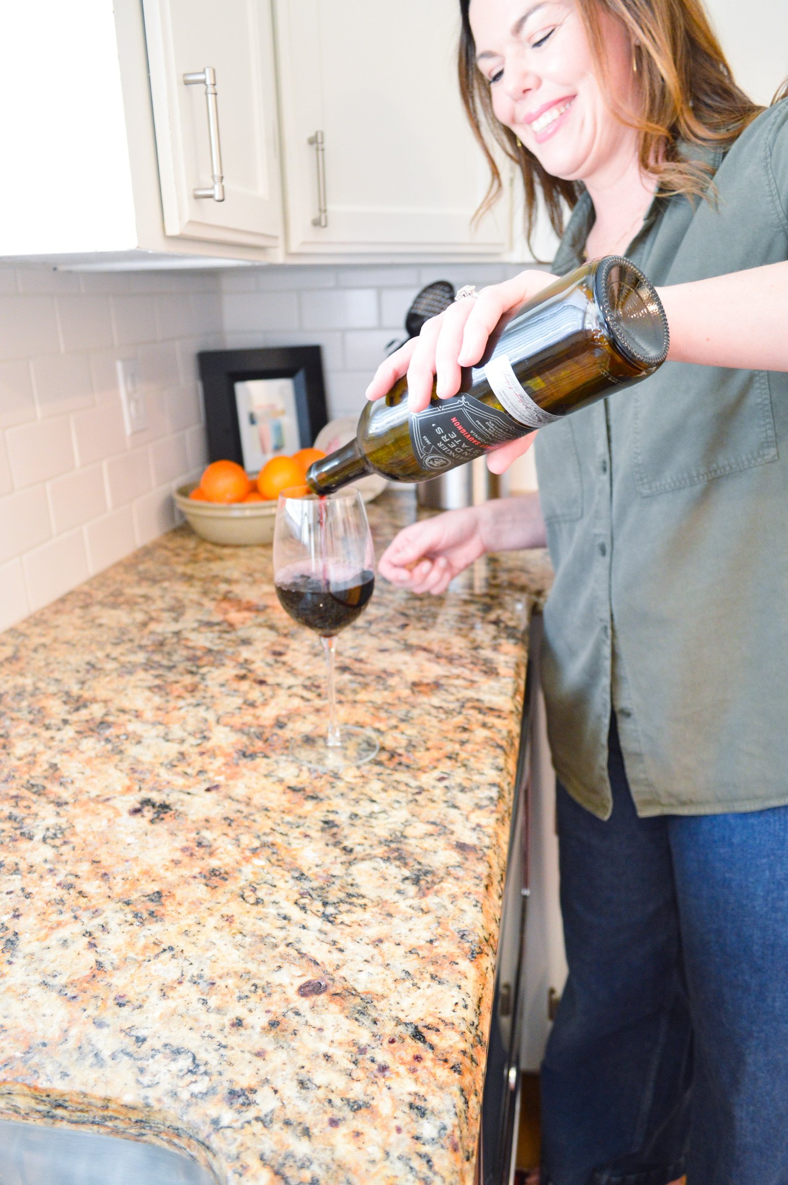 A woman pouring red wine into a wine glass in a kitchen with a granite countertop and a bowl of oranges in the background.