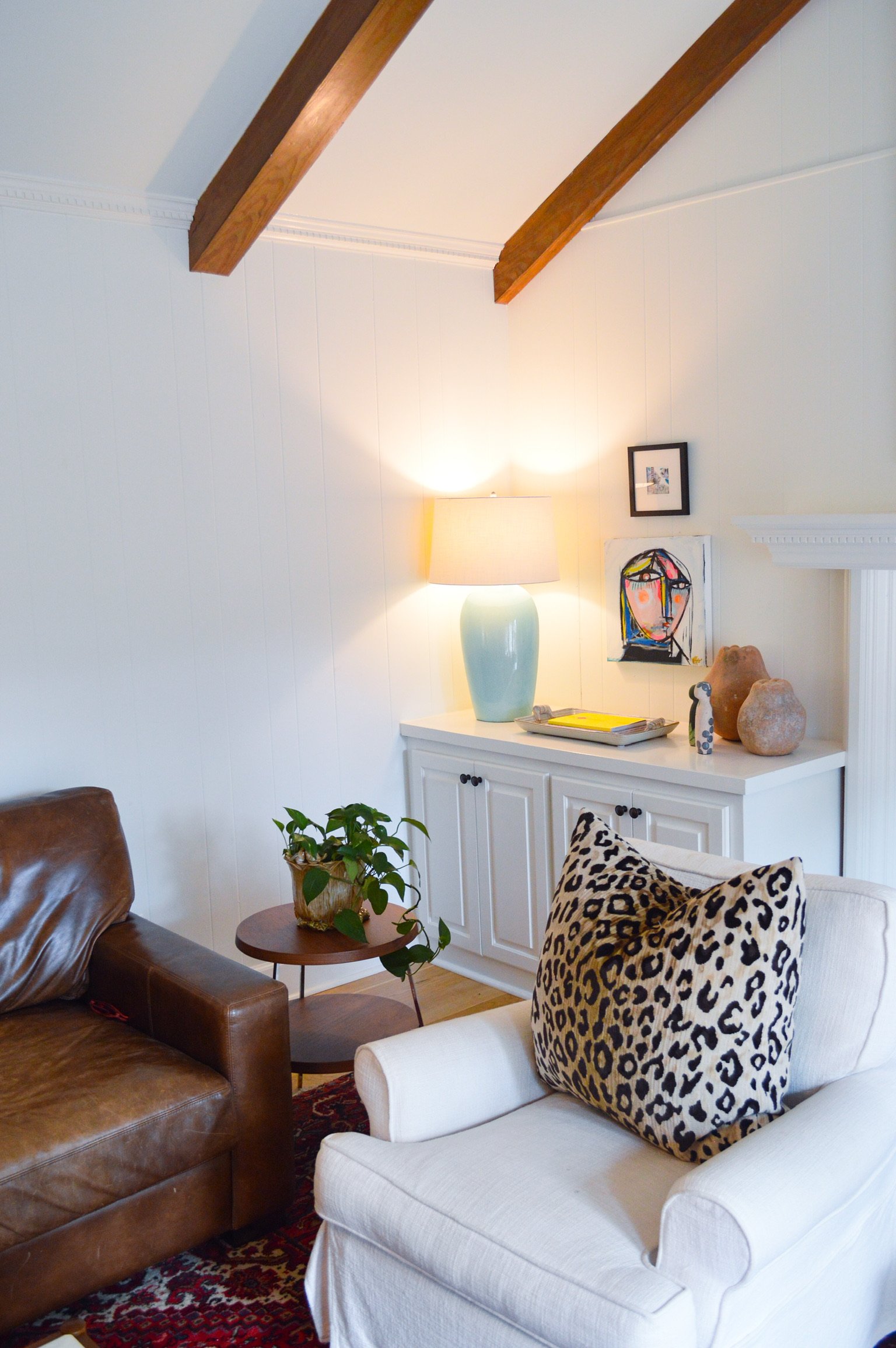 Living room with white walls, exposed wooden beams on the ceiling, a white cabinet with decorative objects, a table with a green plant, a leather armchair, a white armchair with a leopard print pillow, a floor lamp, and artwork on the wall.