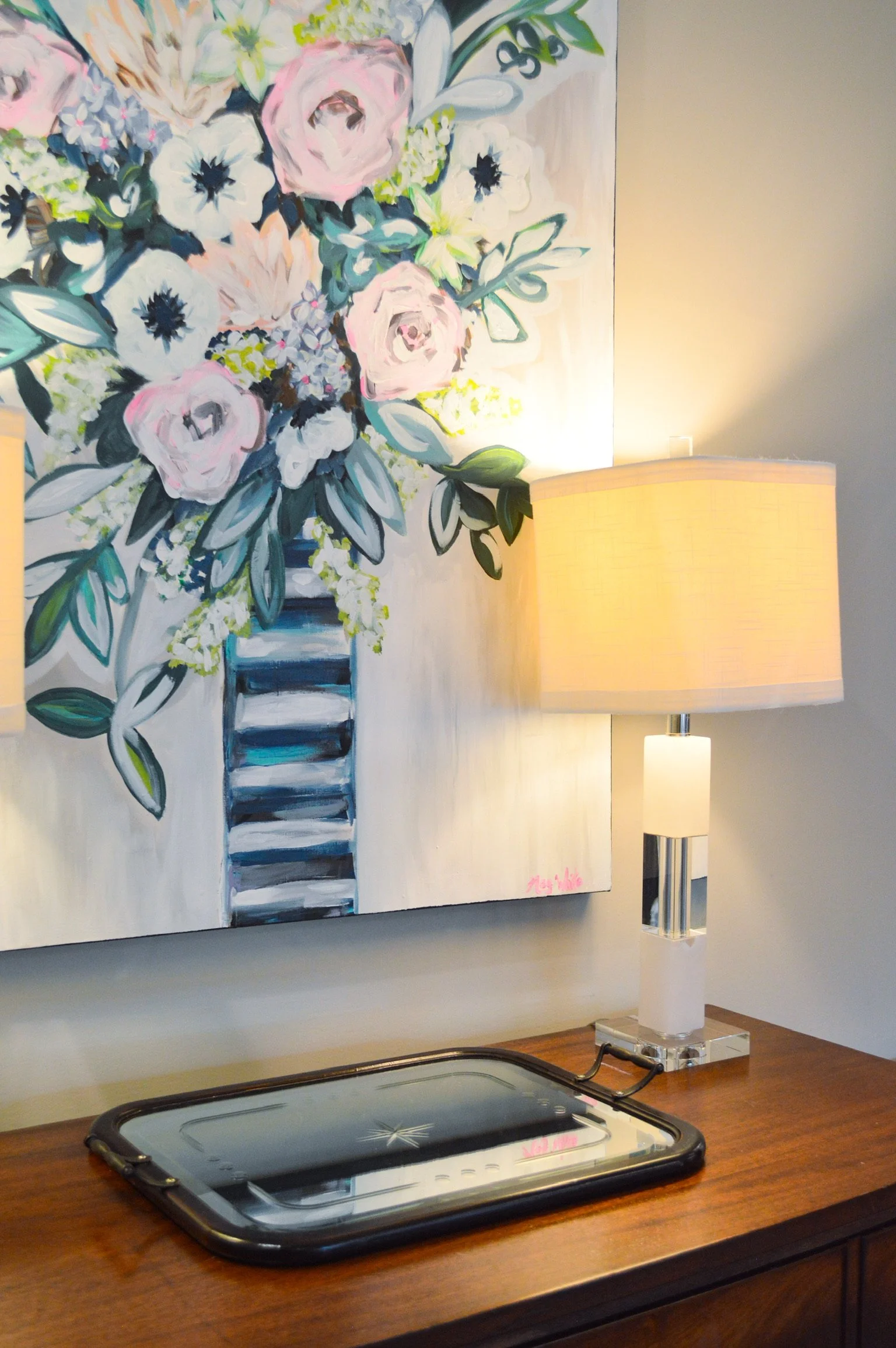 A bouquet of pink and white flowers in a blue and white striped vase on a wooden table, with a beige lamp and a tray nearby, against a wall with a floral painting.