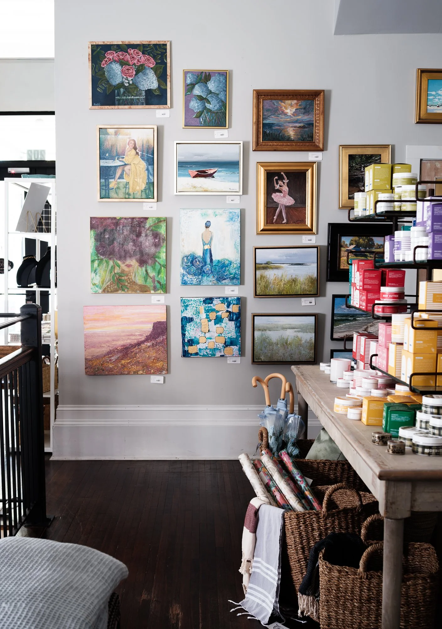 An art gallery displays multiple paintings on a light gray wall, including floral, landscape, and figure artworks. A wooden table holds various art supplies like paint jars and brushes, with woven baskets underneath. In the foreground, an umbrella and a white cloth are visible.