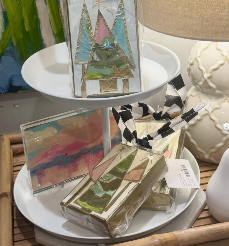 Decorative holiday-themed gift boxes with gold and pastel designs, wrapped with black and white striped ribbons, arranged on a two-tiered white tray near a lamp and a ceramic vase.