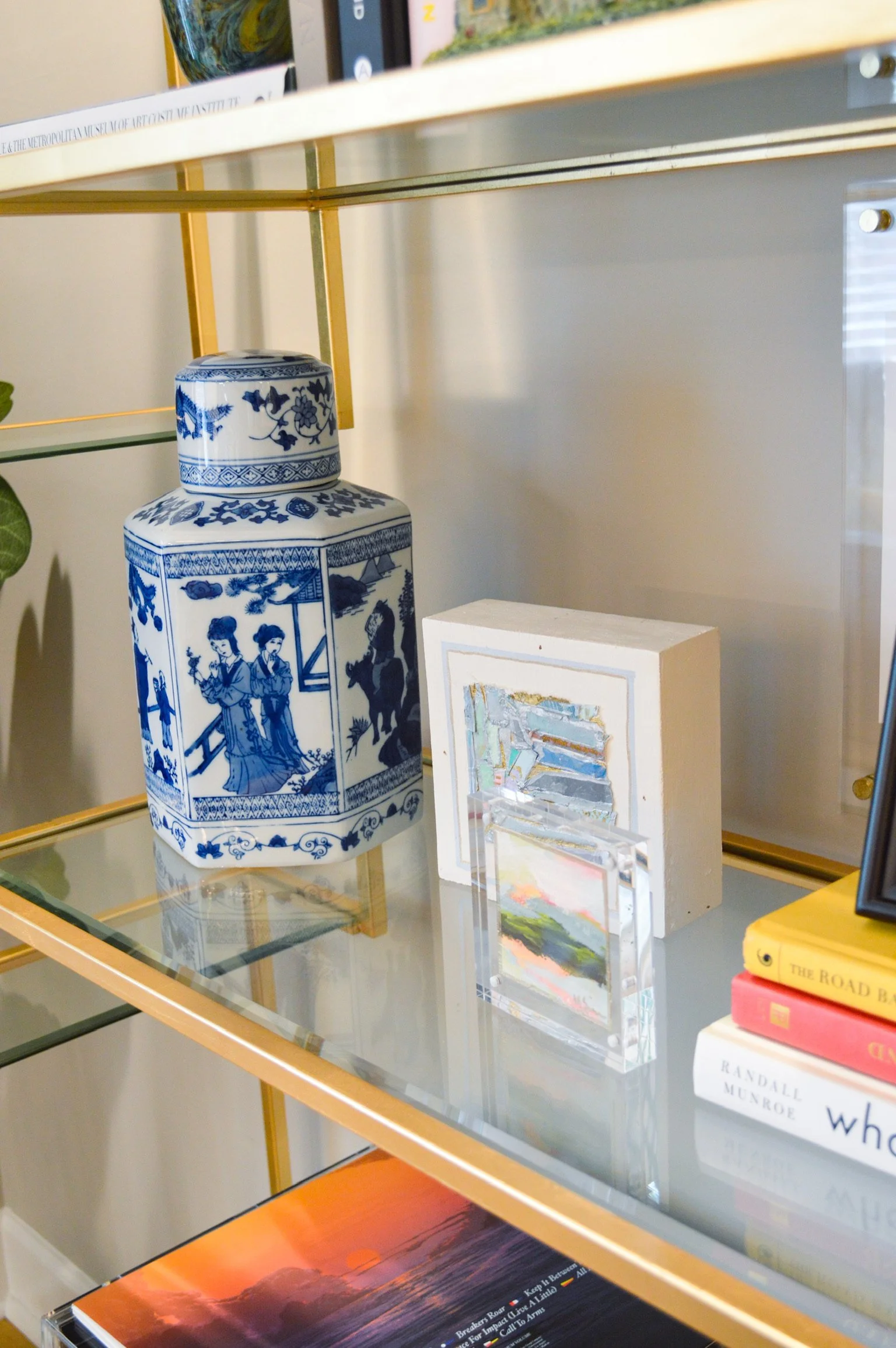 Decorative shelf holding a blue and white porcelain jar, a white framed artwork, small colorful paintings, and books.