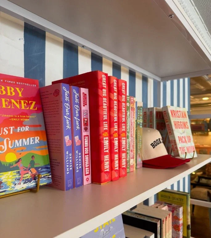 Books on a shelf, including "Just for Summer" by Bobby Bones, multiple copies of "Great Big Beautiful Life" by Emily Henry, a white cap with a red brim and the words "BOOKS" on the front, and a paperback book titled "Kristan Higgins".