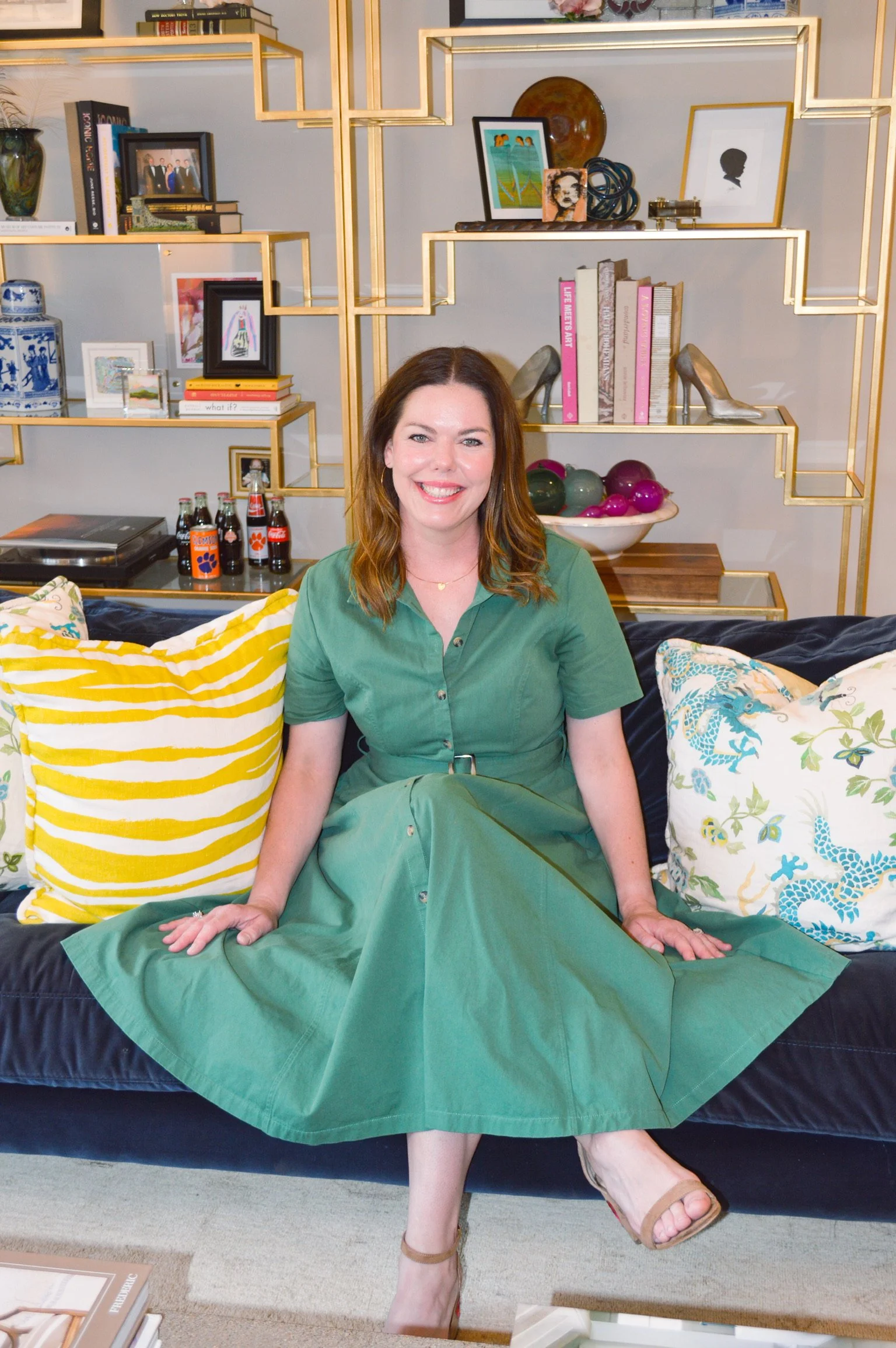A woman with brown hair, wearing a green dress, sitting on a black sofa with colorful pillows, in a modern living room with decorative shelves, artwork, and bottles.